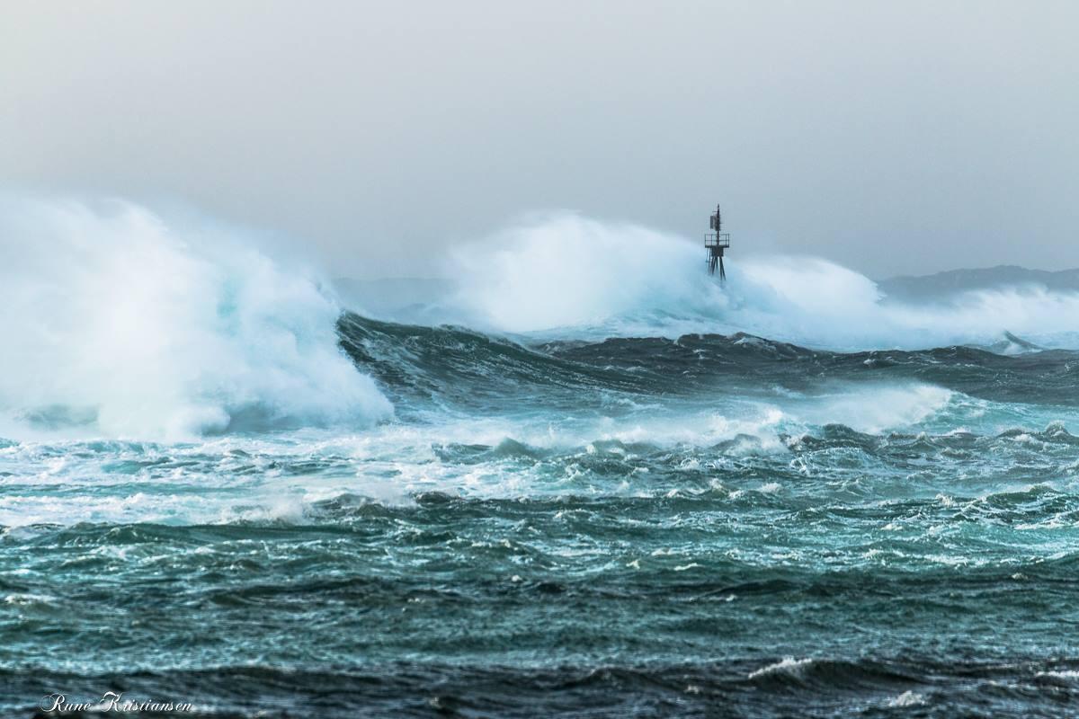 Voldsomme bølger krasjer mot et fyr under en stormfull grå himmel.