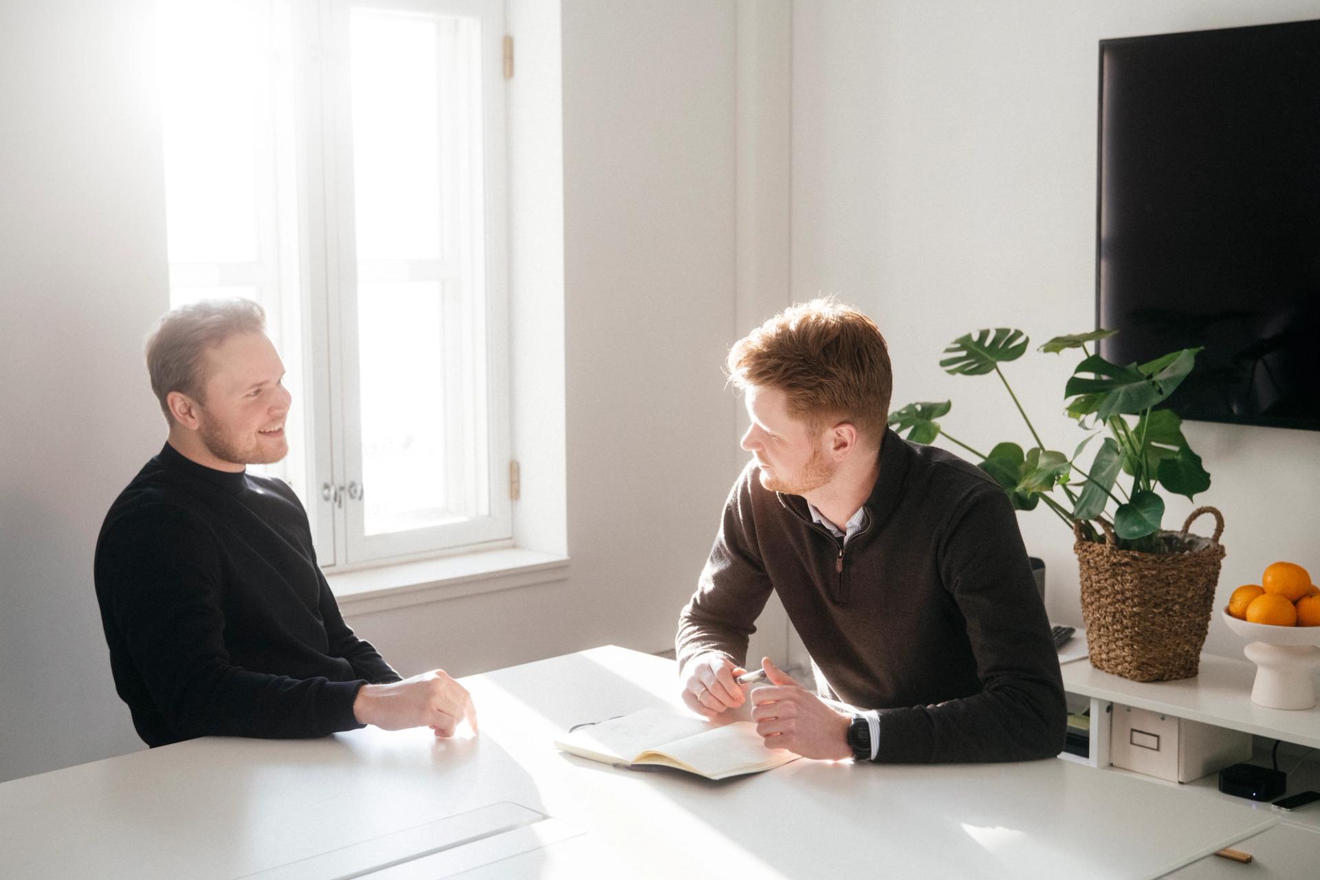 To menn sitter ved et bord og snakker, med sollys som stråler inn fra et vindu.