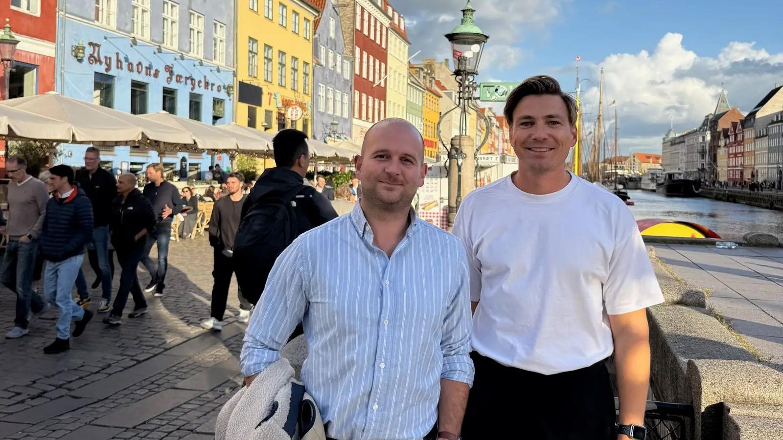 To menn smiler foran kanalen i Nyhavn, København, med fargerike hus og folk i bakgrunnen.
