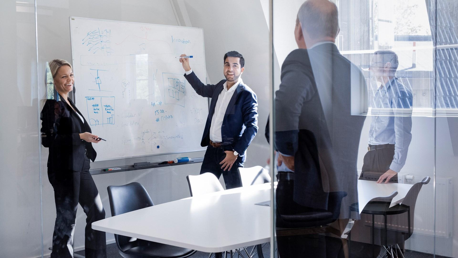 To forretningsfolk presenterer for en kollega i et møterom med en whiteboard full av skriblerier.