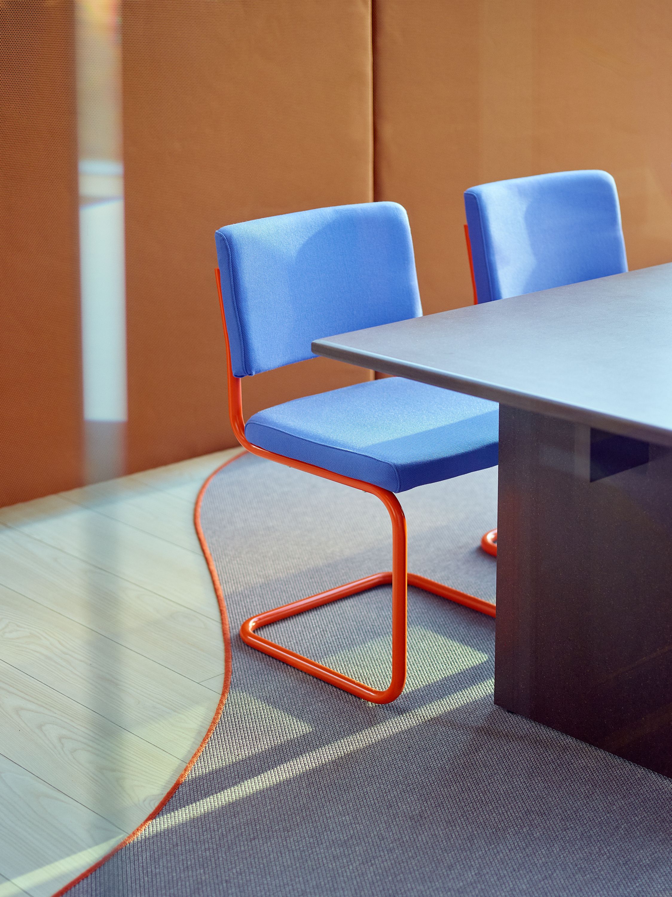 Et moderne møterom med to blå stoler med oransje ramme, et mørkt bord, tregulv og et grått teppe.