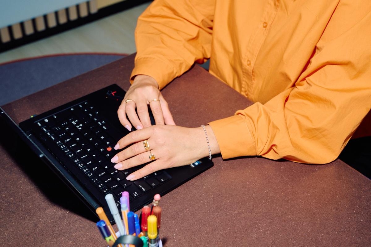 En kvinne med oransje skjorte og smykker skriver på en svart bærbar datamaskin.