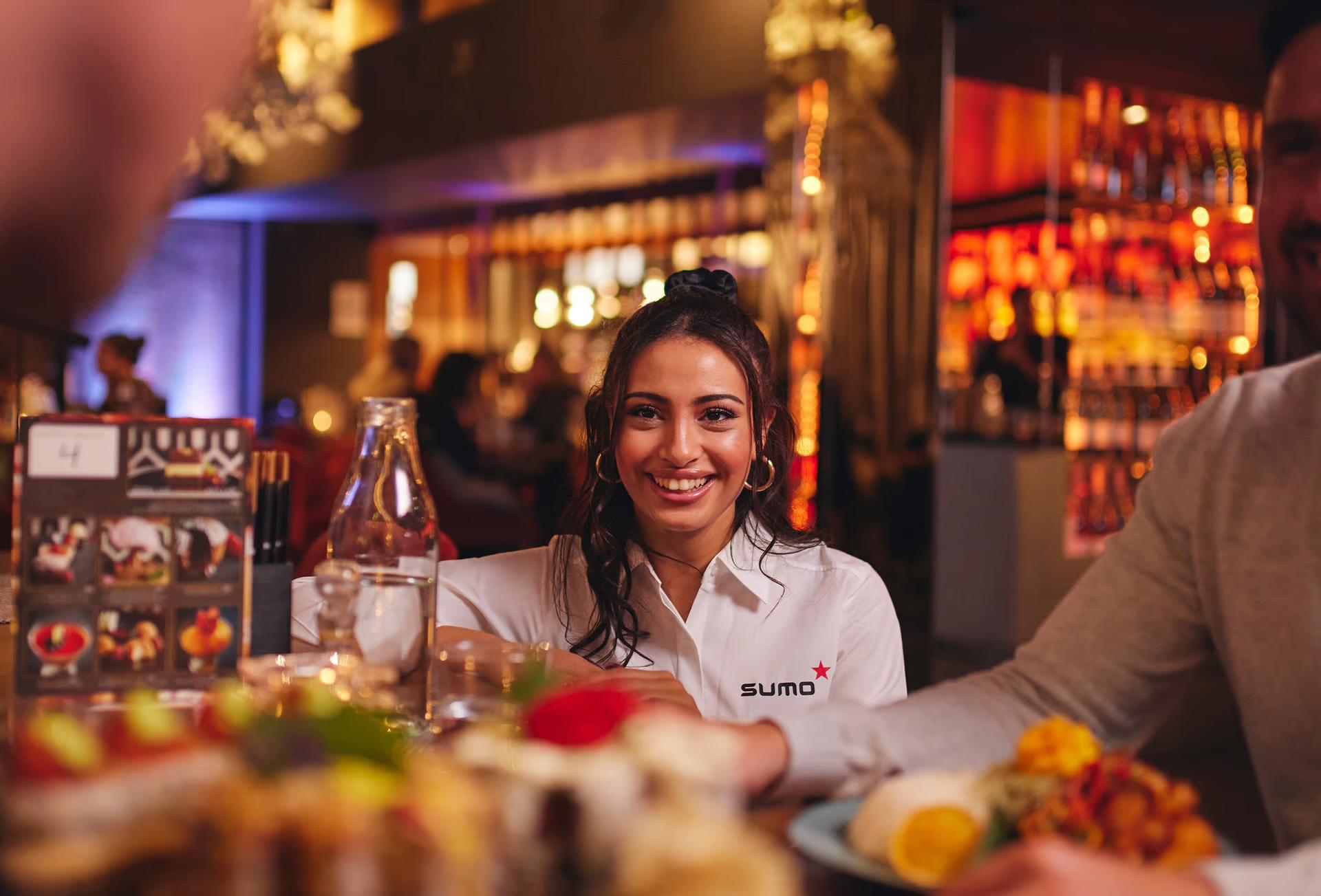 Kvinne i hvit skjorte smiler til kamera ved et restaurantbord.