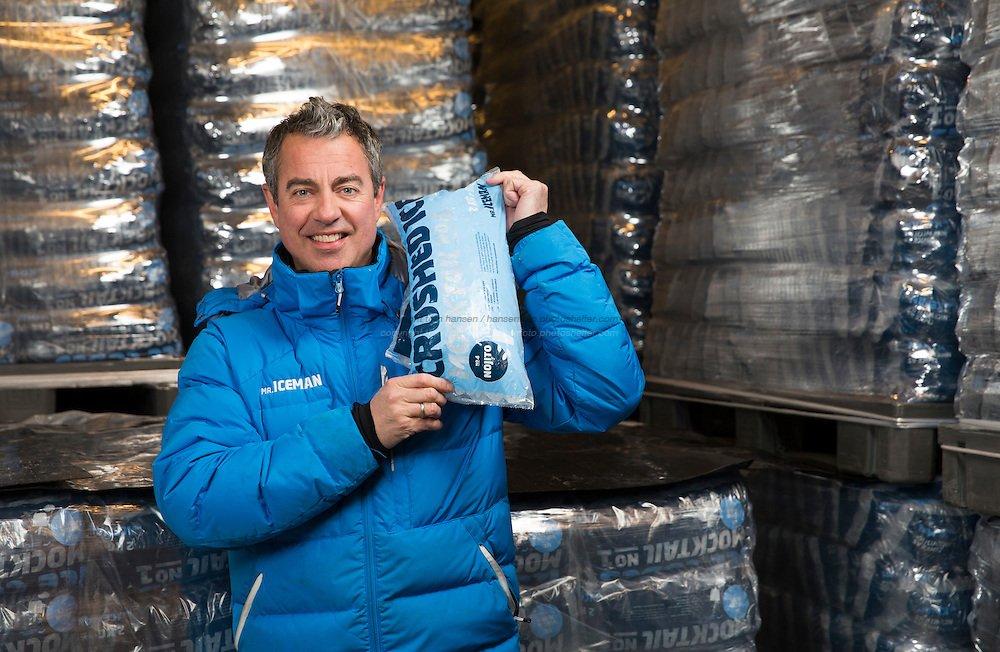 Mann i blå boblejakke smiler og holder opp en pose knust is. I bakgrunnen er stabler med ispakker.