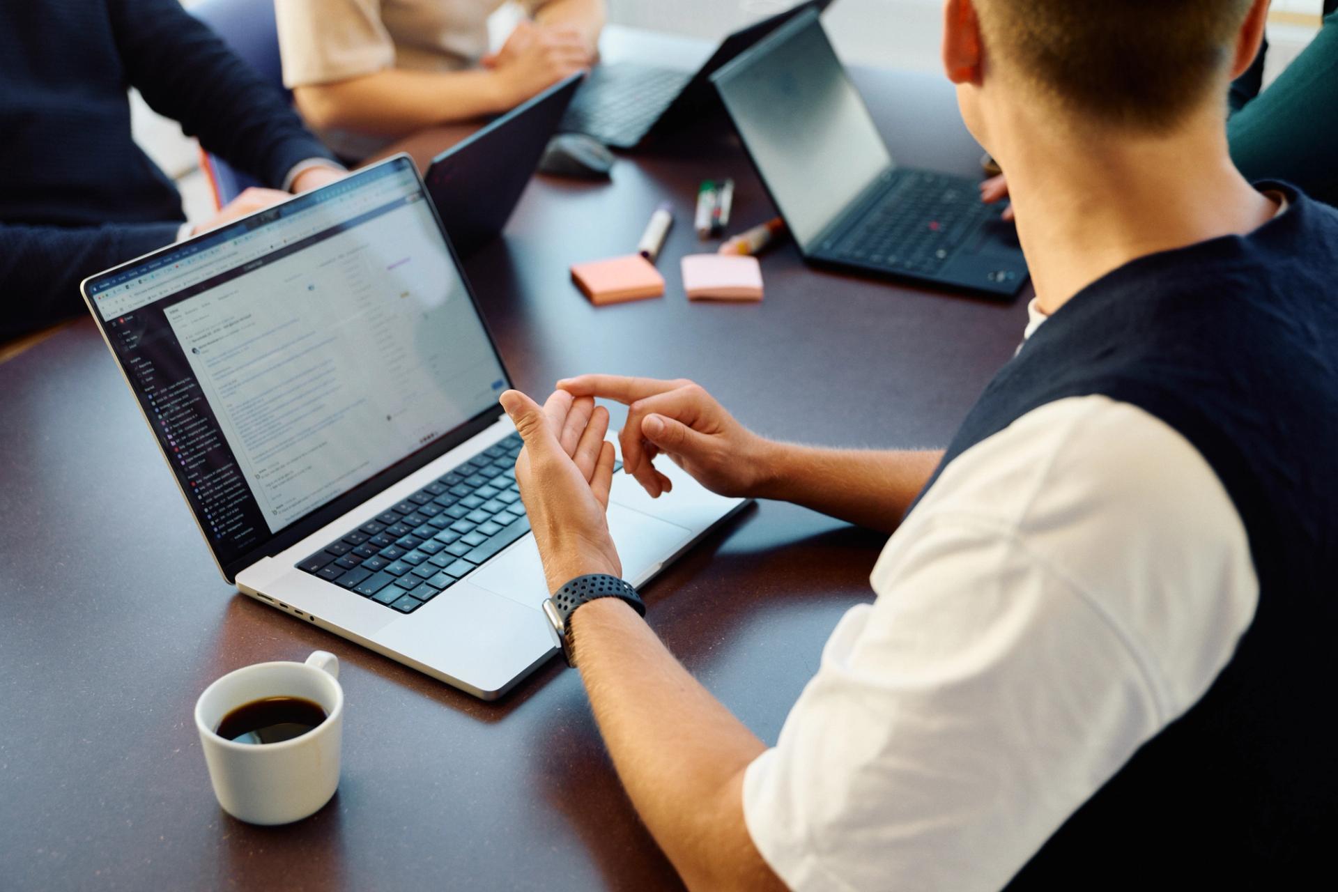 En person med smartklokke berører sin hånd, sitter foran en bærbar PC med kaffe på et kontorbord.