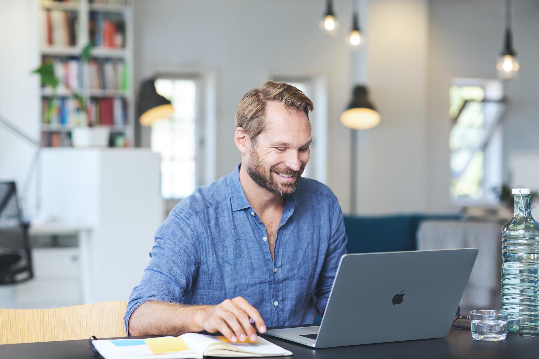 En smilende mann med skjegg og blå skjorte sitter ved et bord med en bærbar PC og notatbok.