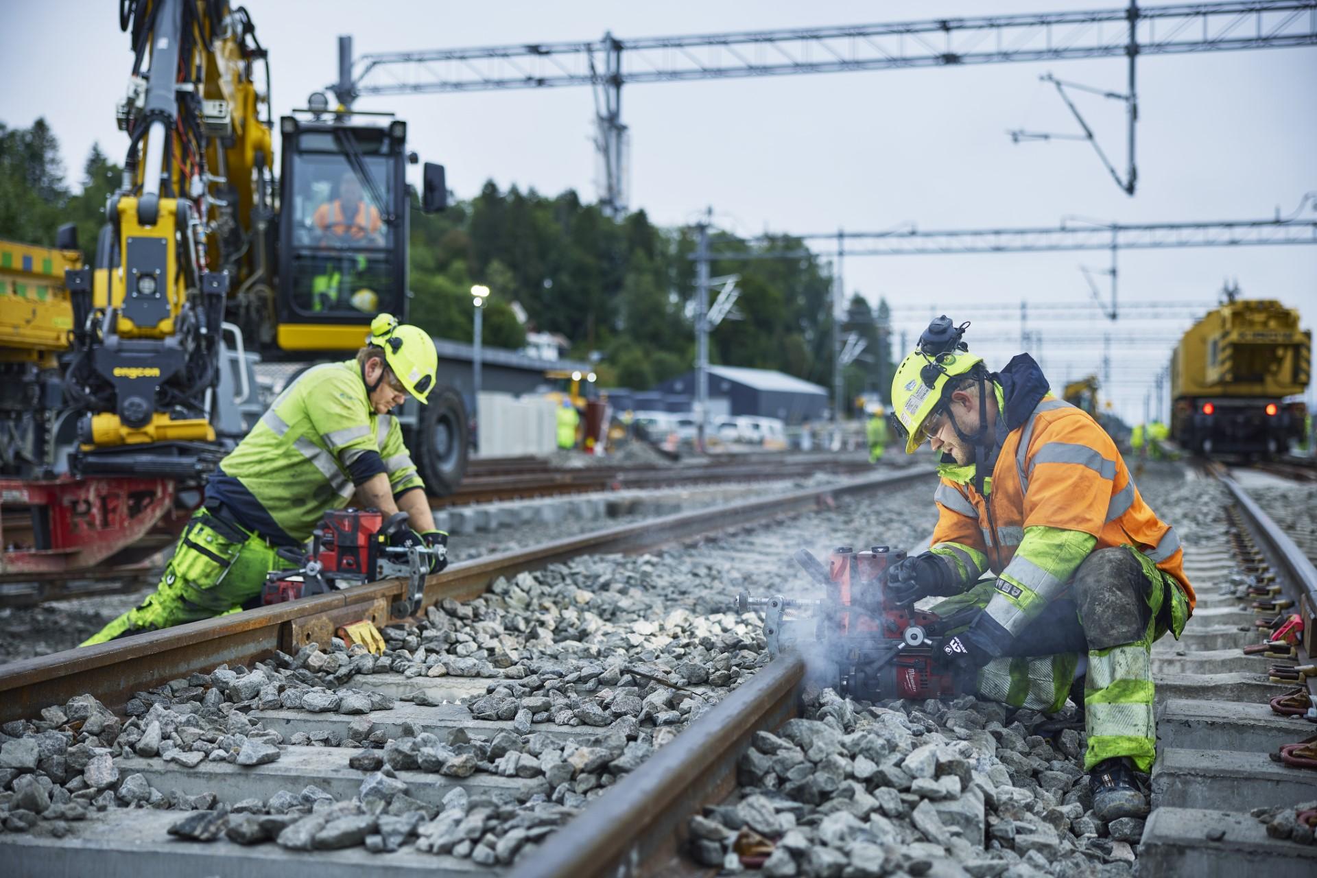 To arbeidere i refleksvester og hjelm borer i togskinner mens anleggsmaskiner arbeider i bakgrunnen.