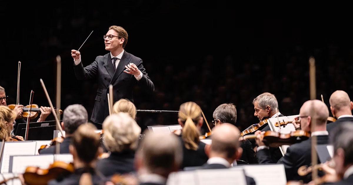 En dirigent leder et orkester i en konsertsal, sett bakfra publikum og musikerne.
