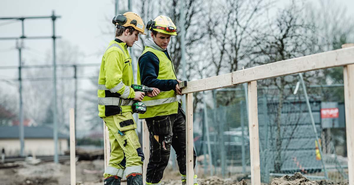 To byggearbeidere i gul refleksvest og hjelm monterer treplater med drill på en byggeplass.