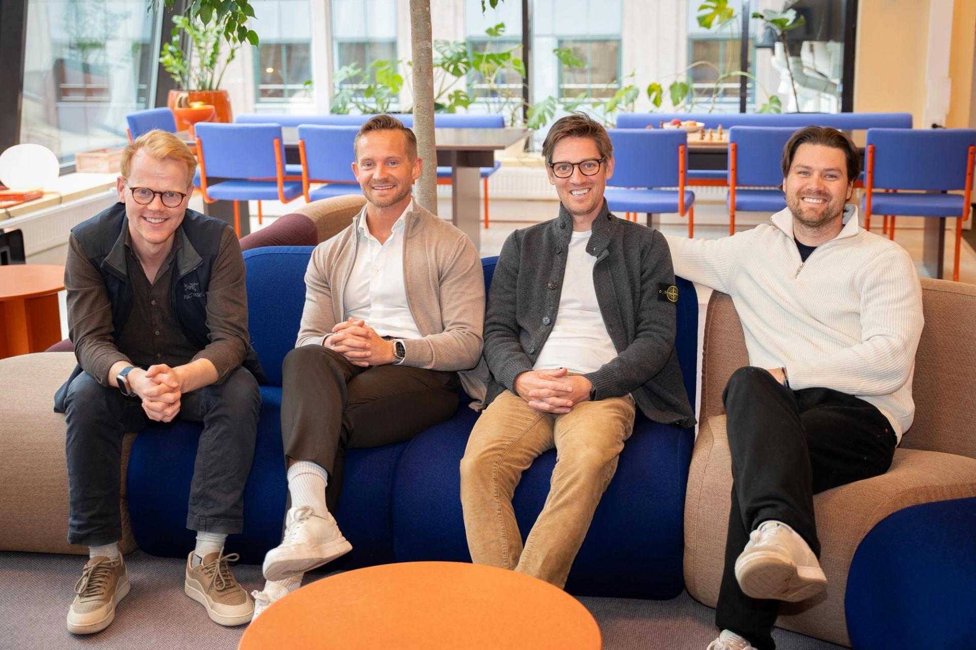 Fire menn sitter i en sofa på et lyst kontor med blå stoler og planter i bakgrunnen