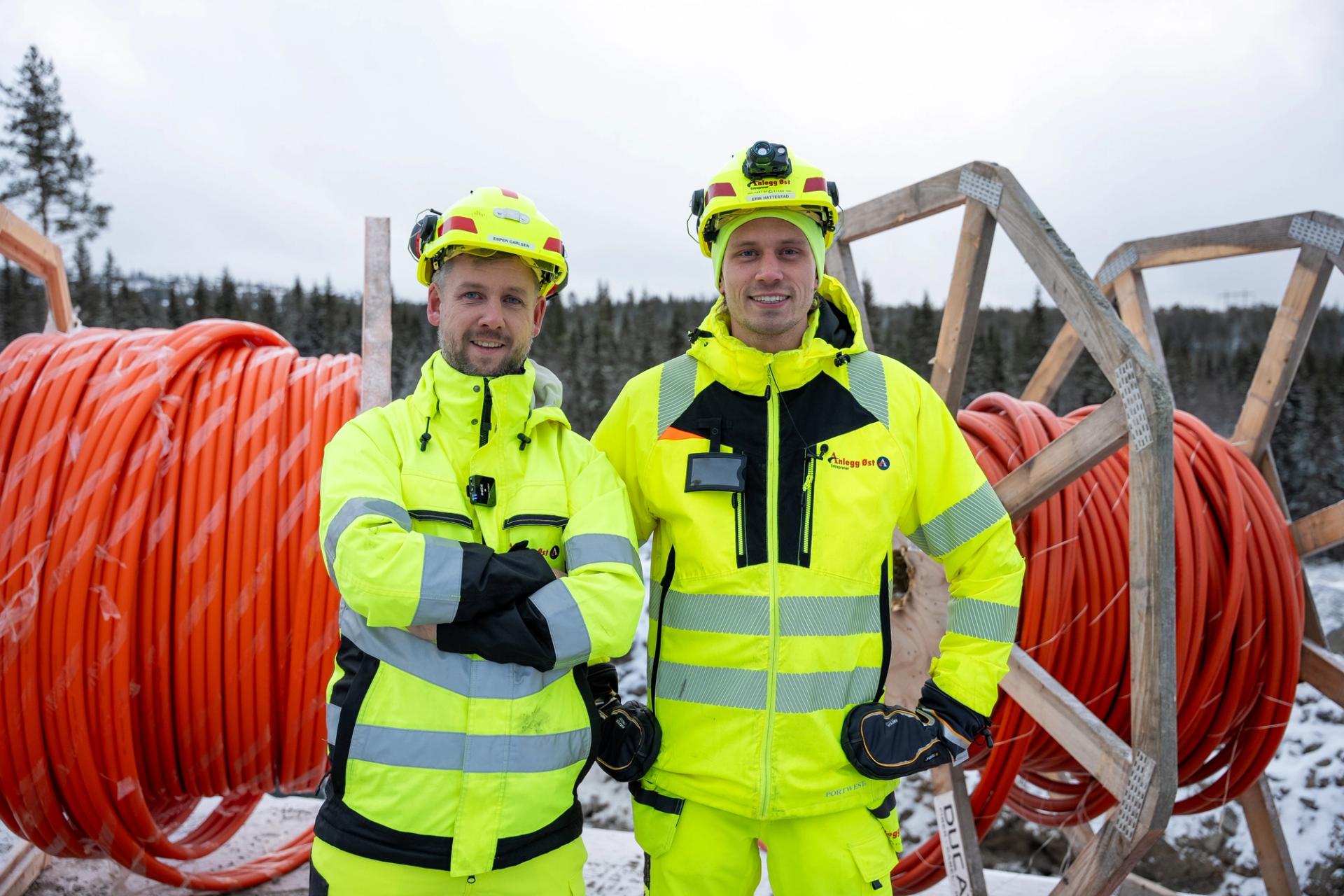 To menn i gule arbeidsklær og hjelmer står smilende foran store kabelruller i et snødekket landskap.