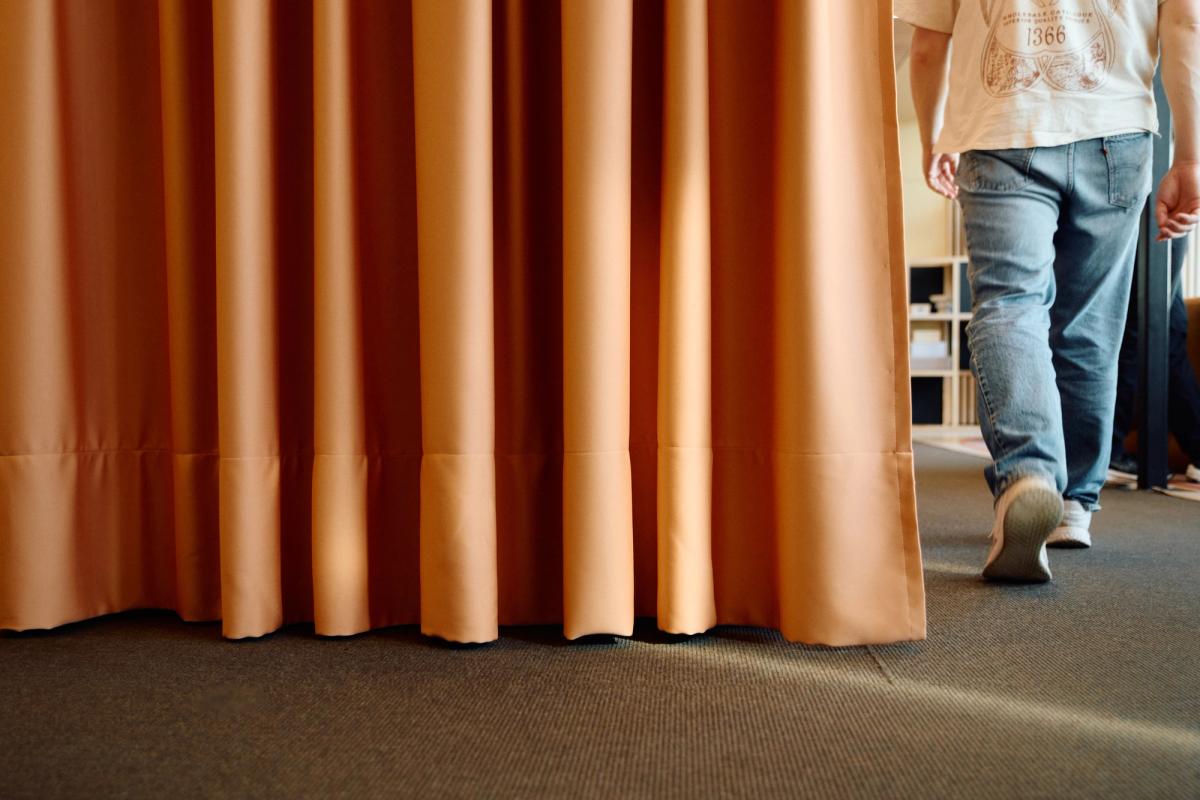 En person i jeans og hvit t-skjorte går inn i et rom, sett bakfra, med et oransje forheng i forgrunnen.