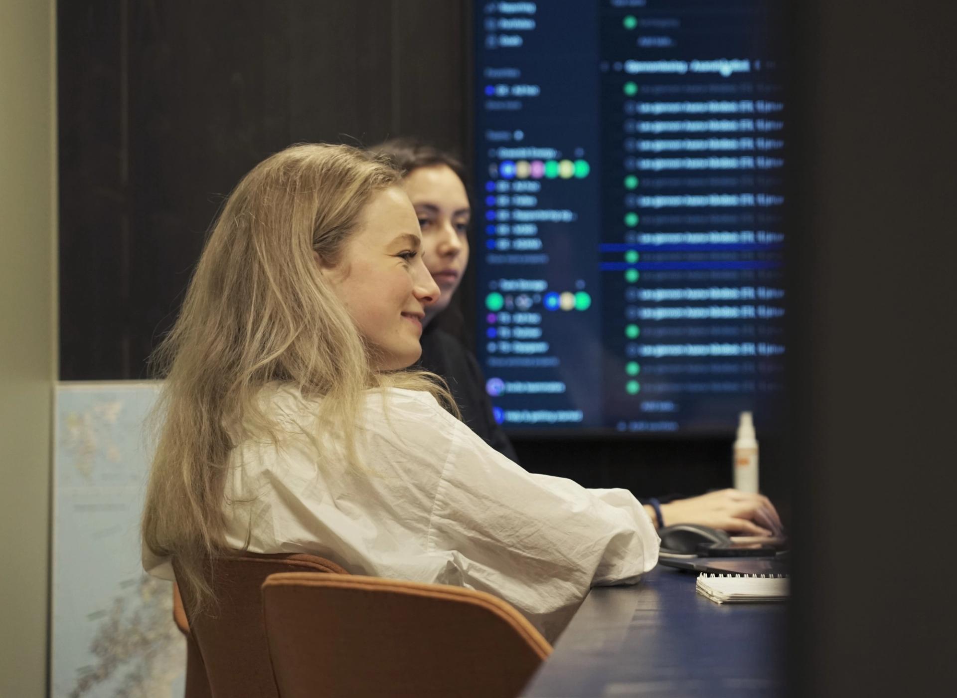 To kvinner sitter foran en stor skjerm med tekst og grønne prikker, en av dem bruker en datamus.