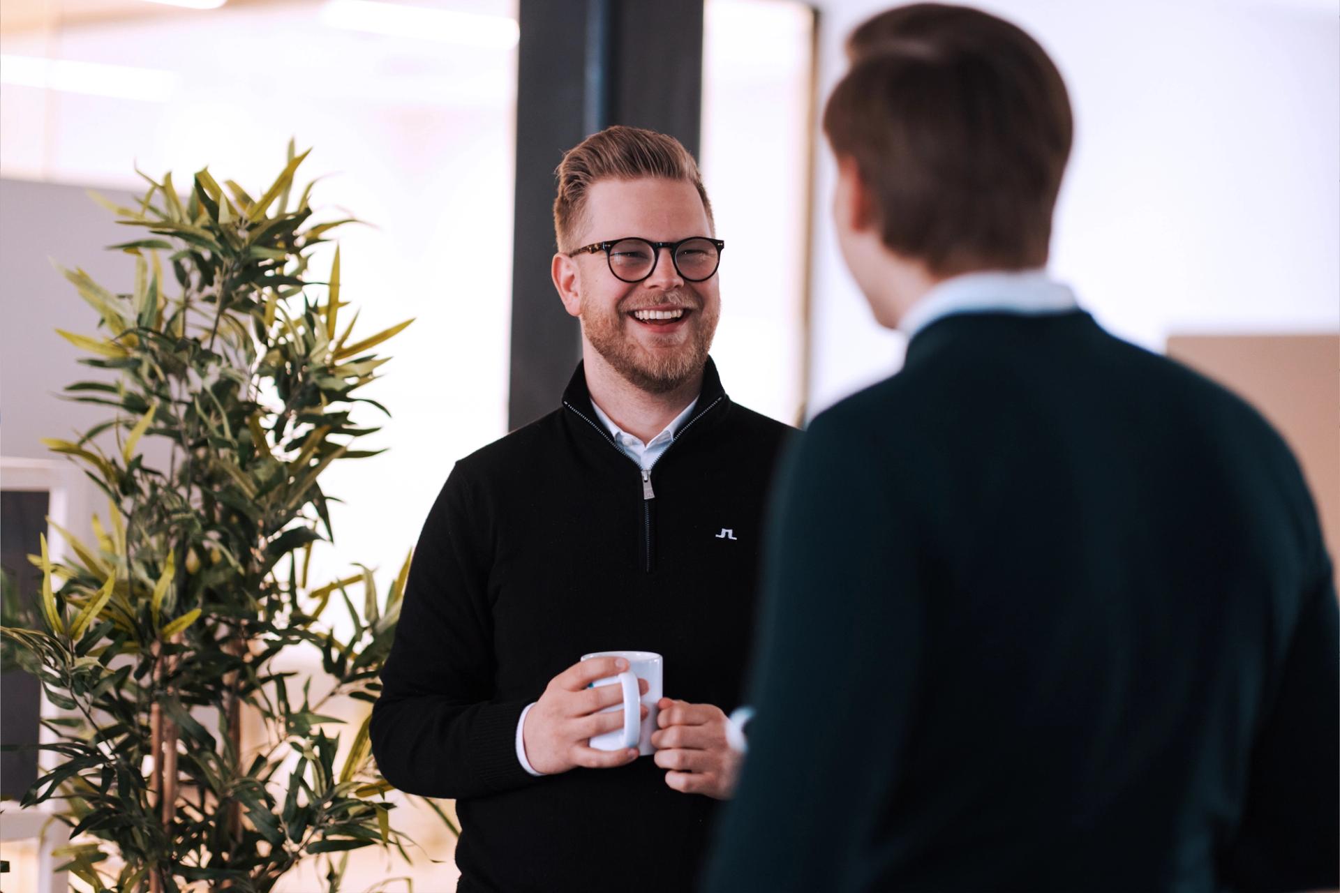 En smilende mann i svart genser holder en kaffekopp og snakker med en person med ryggen til.