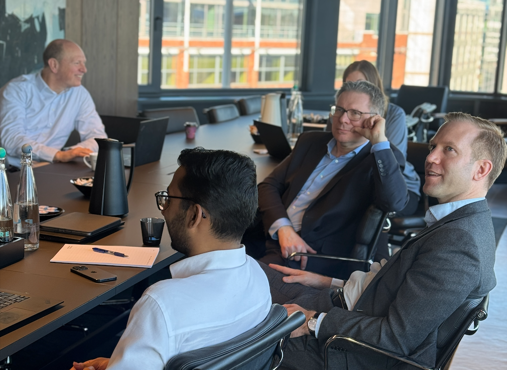Menn i dress og en kvinne sitter rundt et møtebord og diskuterer.