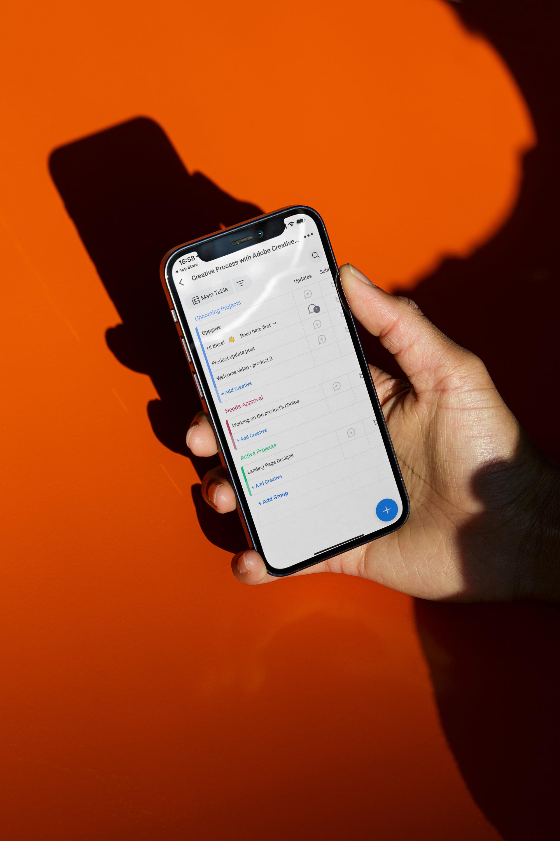 En hånd holder en smarttelefon som viser en prosjektstyringsapp på en oransje bakgrunn.