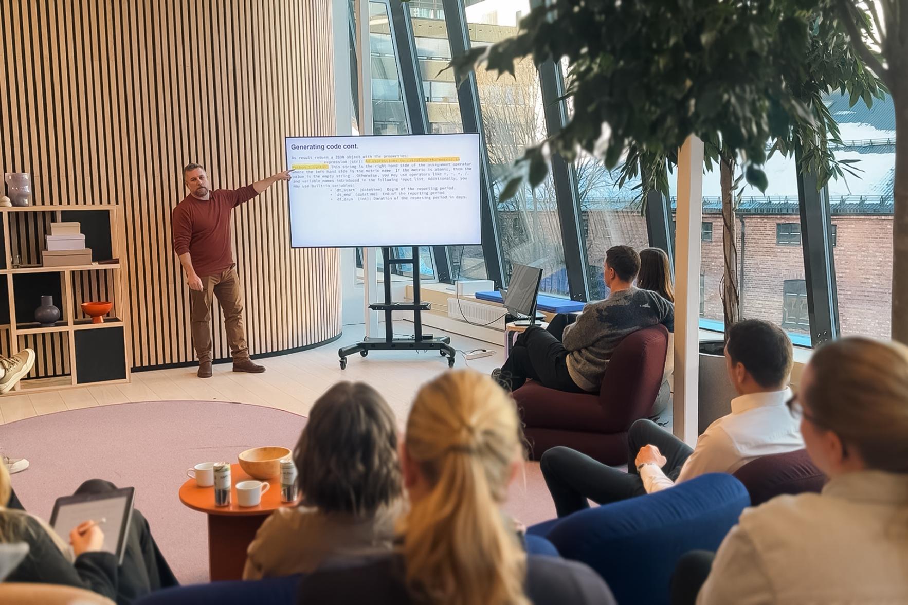 Mann presenterer på seminar om AI-assistenter, med deltakere som lytter i en moderne bygning med store vinduer og planter.