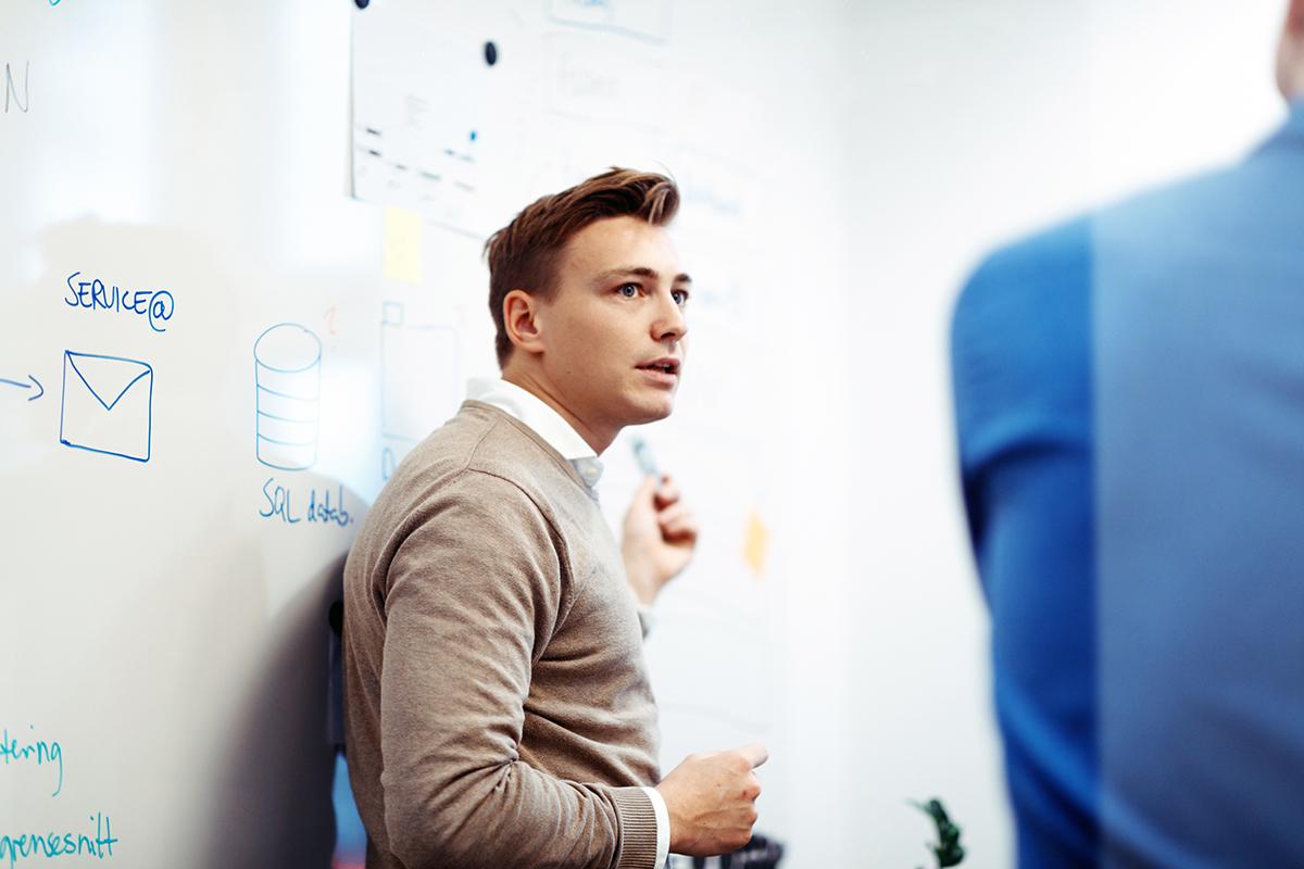 En mann tegner en teknisk diagram på en tavle mens en annen person observerer.