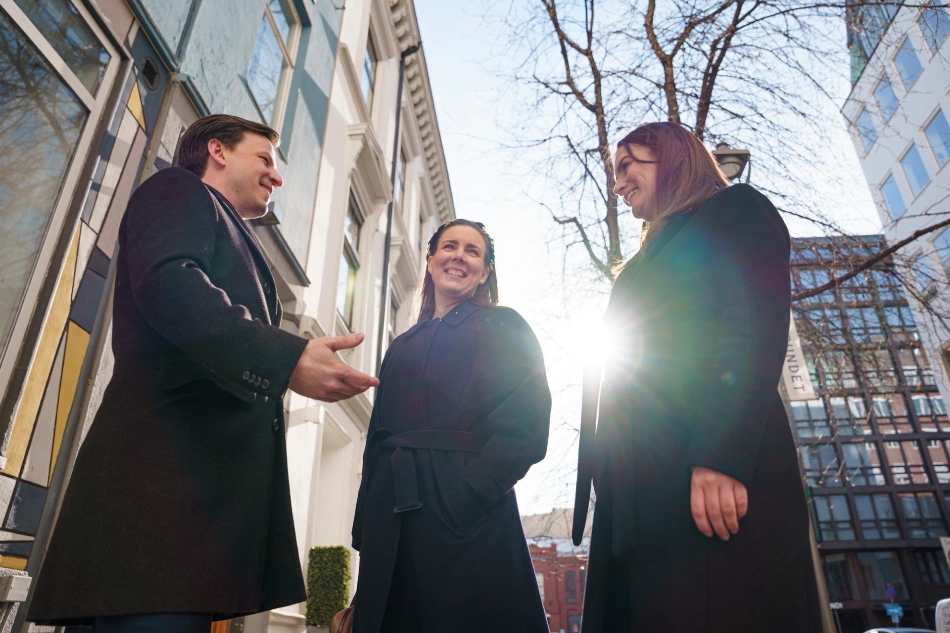 Tre personer i mørke frakker står ute og smiler i solskinnet, med bygninger og nakne trær i bakgrunnen.