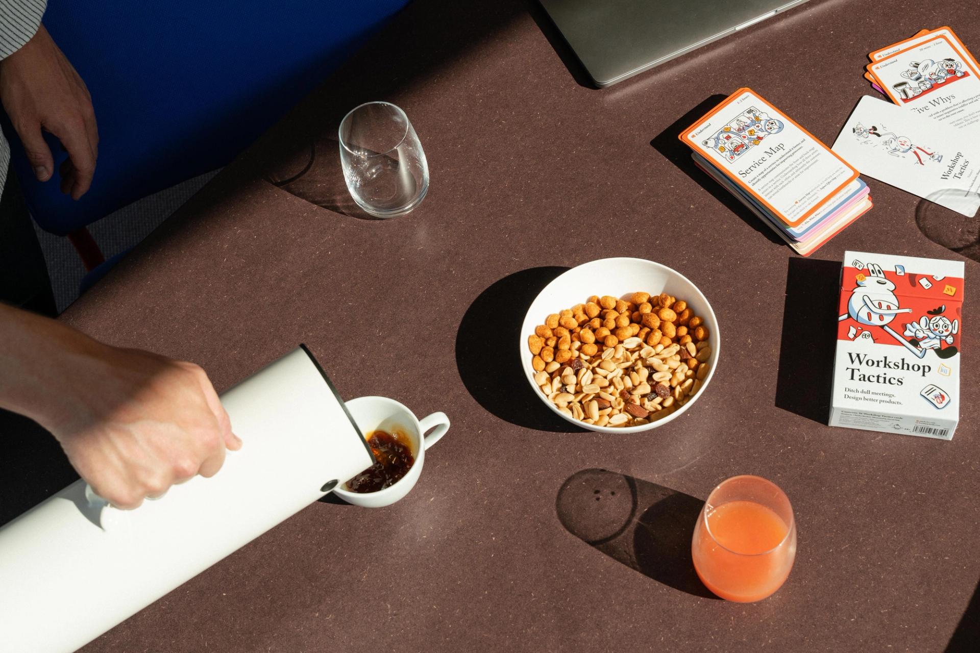 Person heller kaffe, med snacksskål, drikke og kortspill ("Workshop Tactics") på et brunt bord i sterkt sollys.