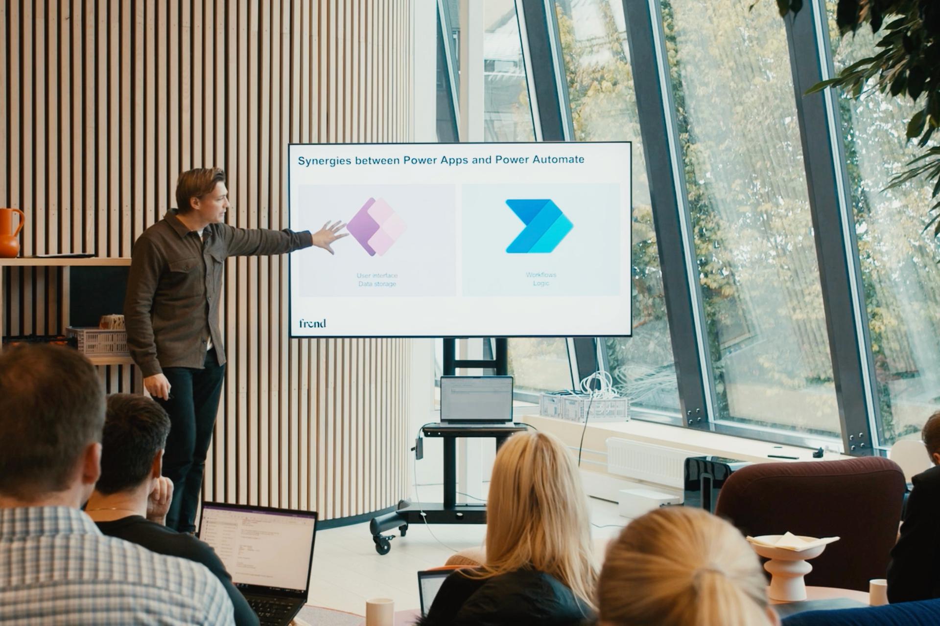 En foredragsholder peker på en skjerm som viser "Synergies between Power Apps and Power Automate" foran et publikum.