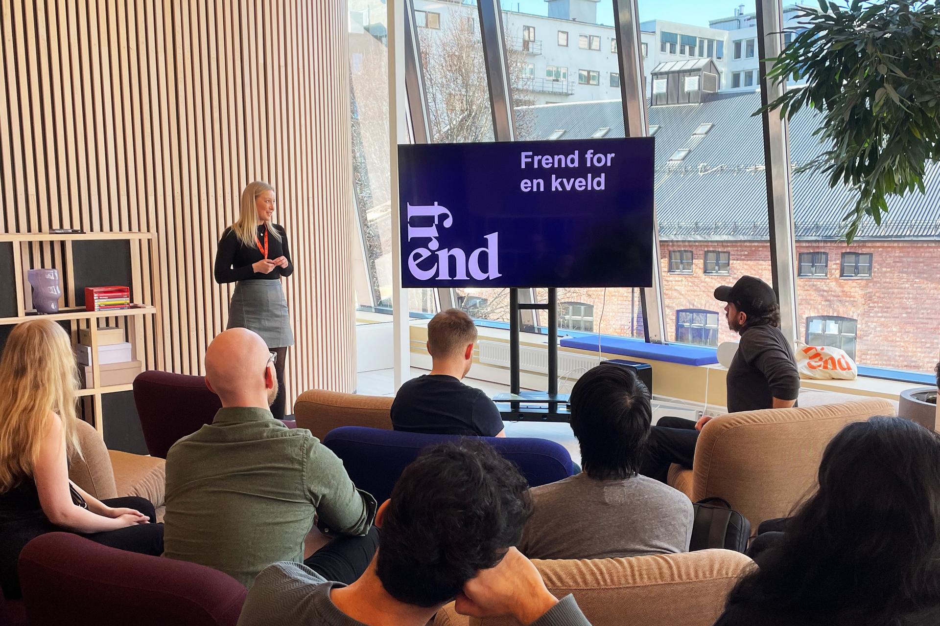 En kvinne presenterer foran et publikum som sitter i sofaer i et rom med store vinduer.