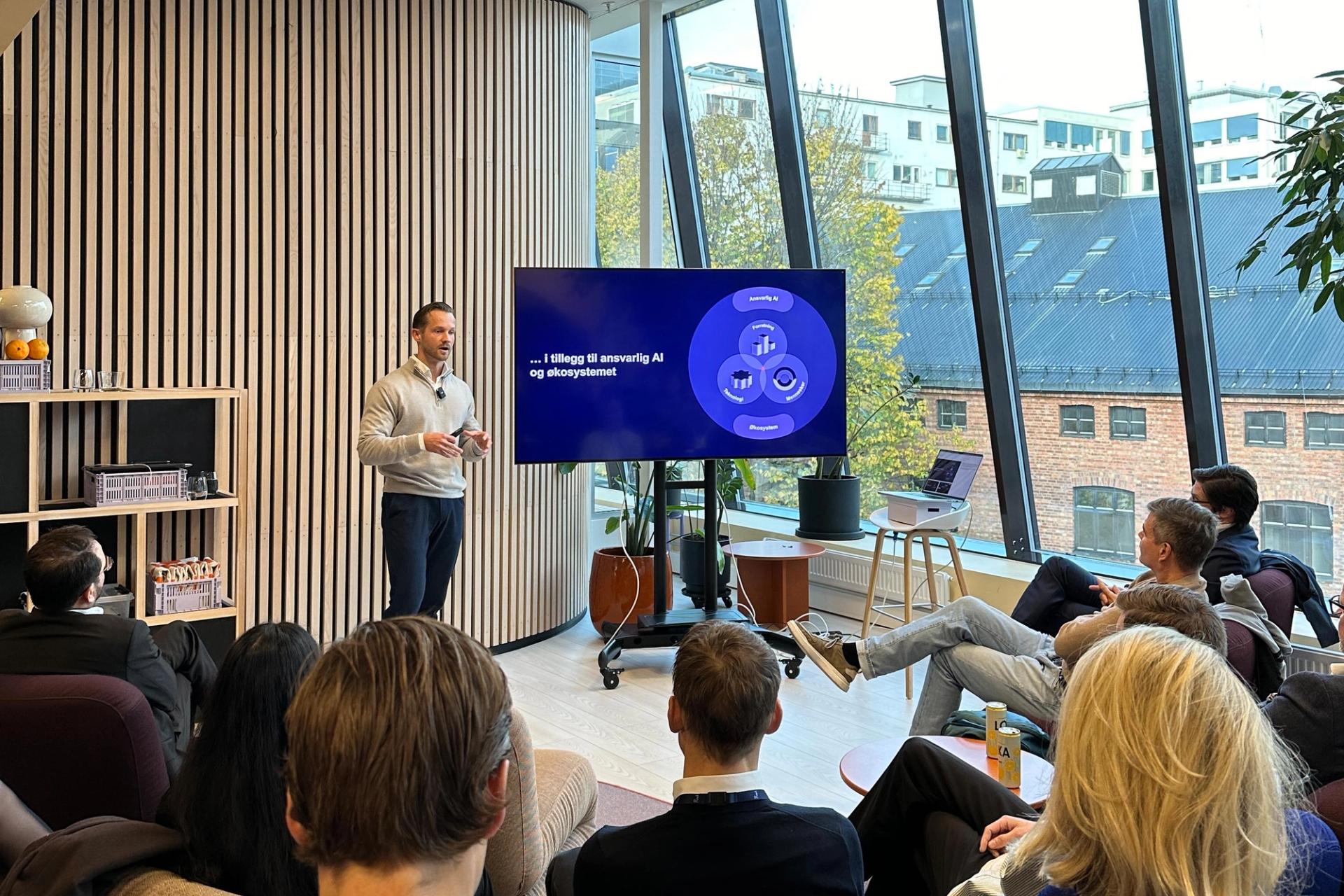 En mann presenterer foran et publikum på et seminar om AI, med et diagram på en skjerm og utsikt over byen.