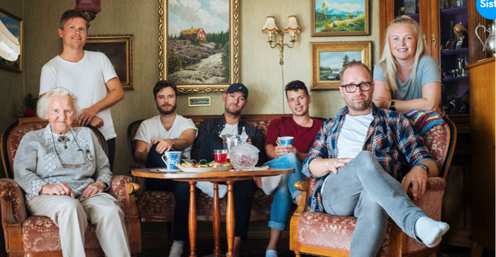 En eldre kvinne og flere yngre voksne sitter og står i en koselig stue, omgitt av malerier og antikke møbler.