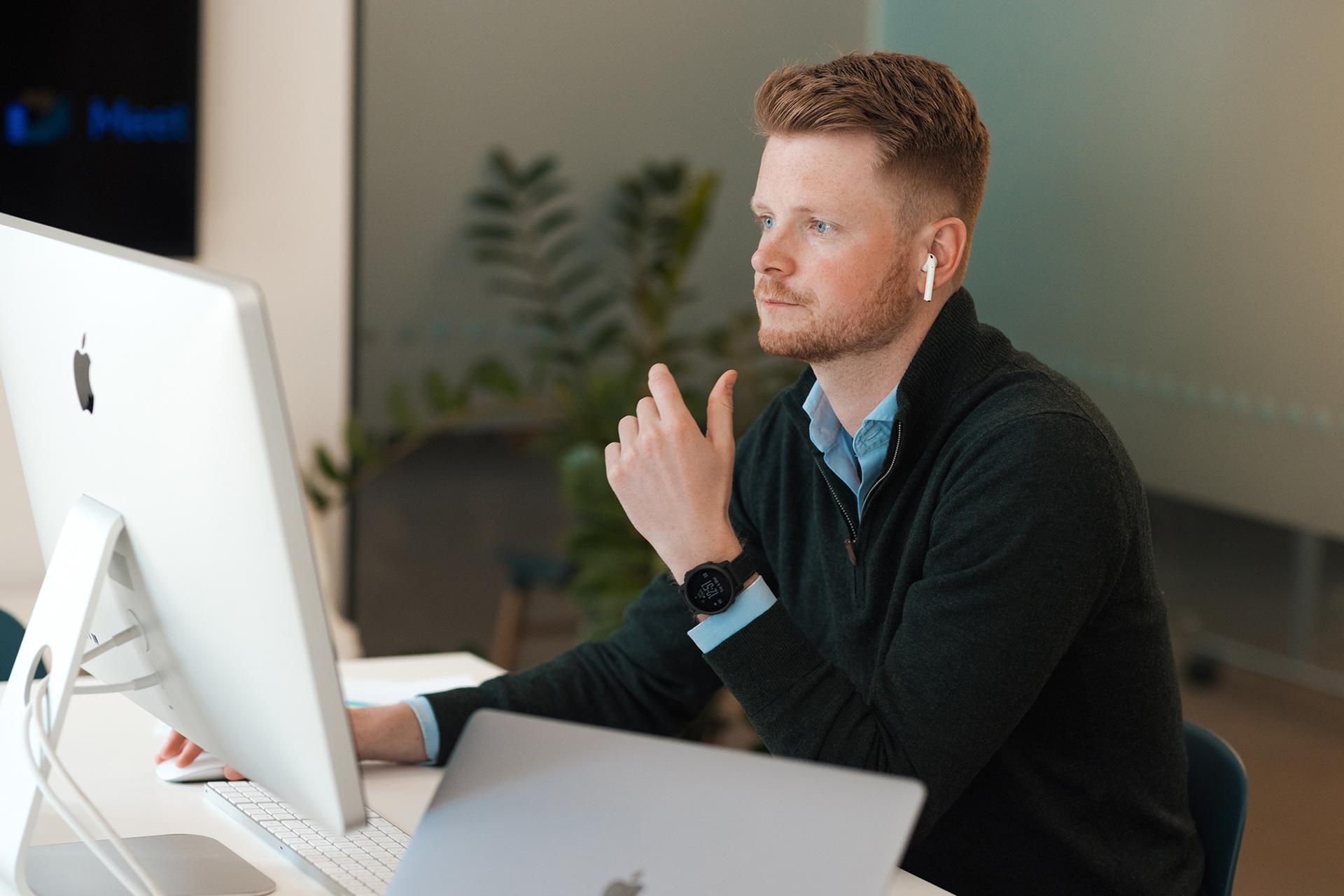 En mann med skjegg og ørepropp sitter ved et skrivebord med Mac-datamaskiner, ser konsentrert ut.