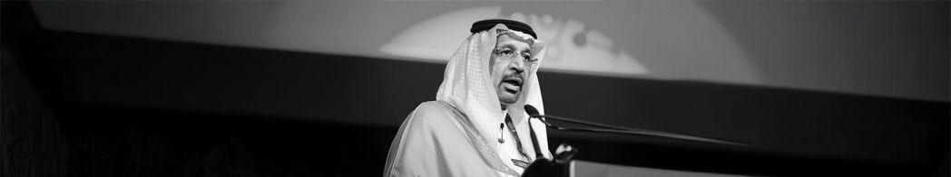 Black-and-white photograph of a speaker in traditional Saudi dress at a podium on stage.