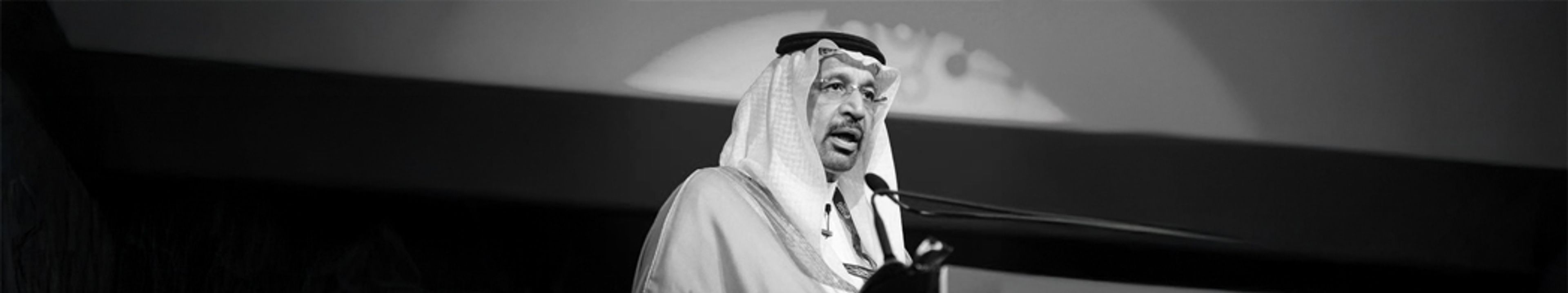 Black-and-white photograph of a speaker in traditional Saudi dress at a podium on stage.