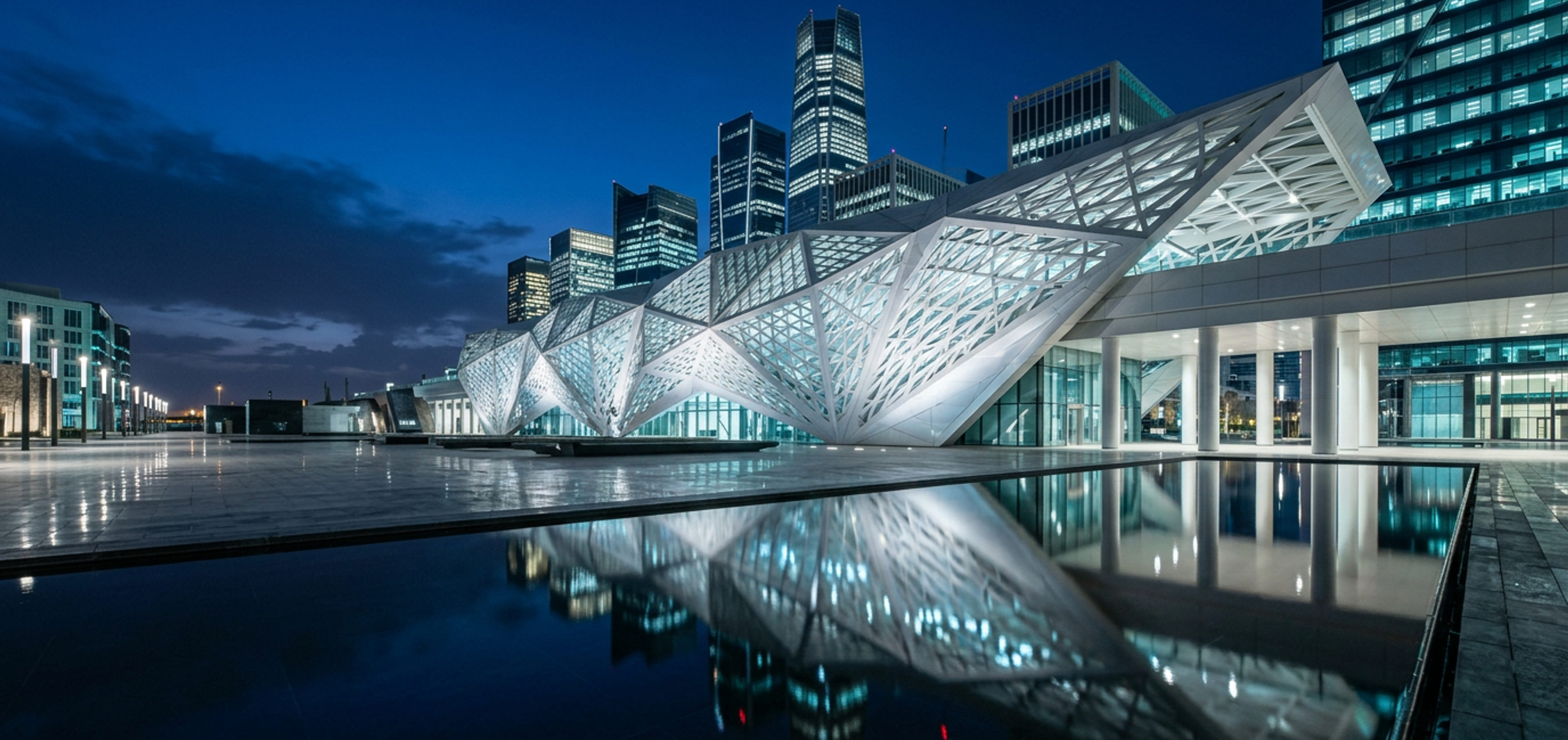 The faceted white lattice of the King Abdullah Financial District metro station reflected in a plaza pool at blue hour.