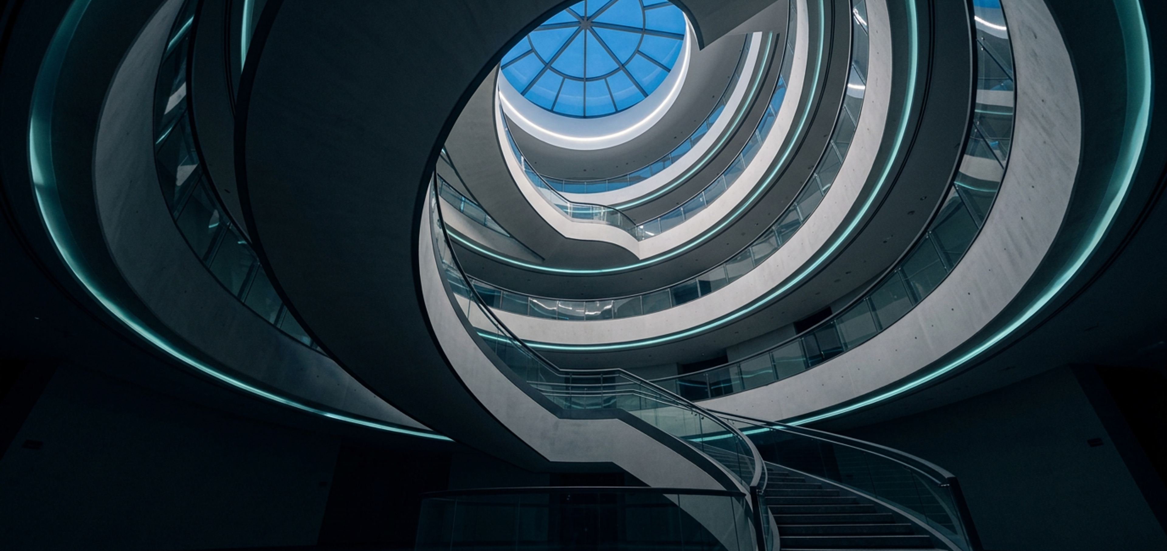 Looking up through a sweeping spiral staircase toward a round skylight in a contemporary Riyadh building.