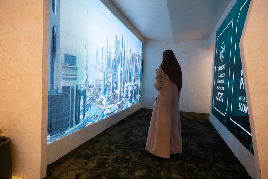 A woman stands in a darkened exhibition room, facing a wall-sized projection of a futuristic city skyline.