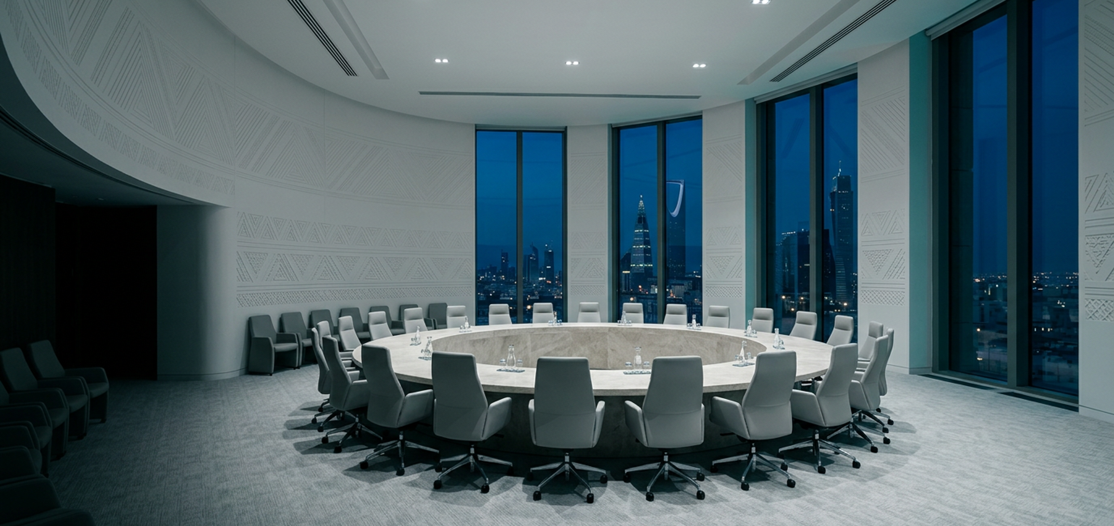 A formal council chamber in Riyadh at blue hour, grey chairs around a round stone table beneath walls carved with Najdi geometric relief.