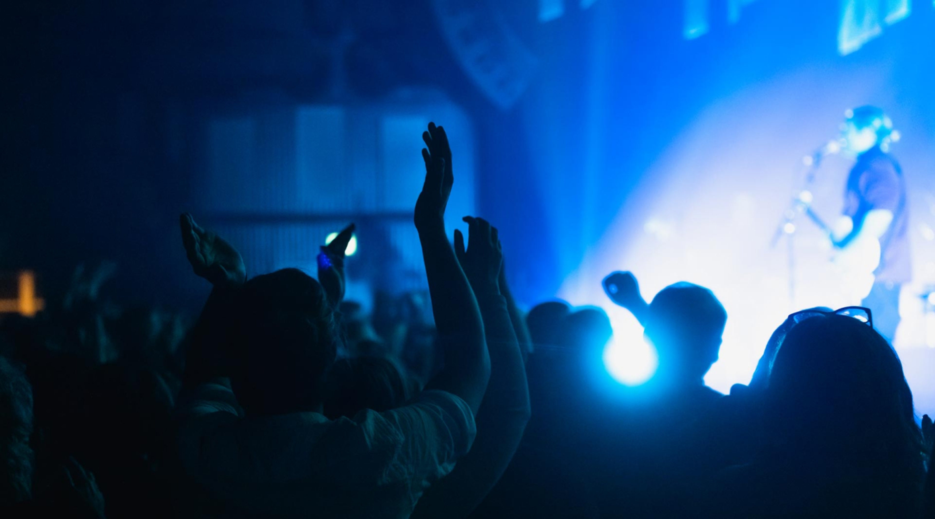 Personer som står i ett publikhav framför en scen med händerna i luften.