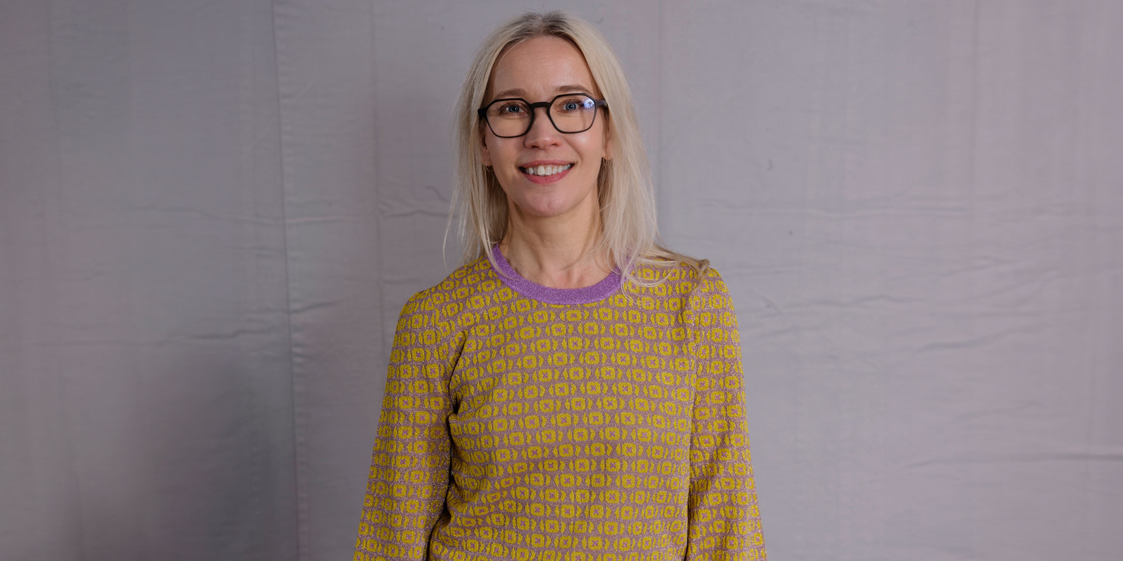 Minna Krook står framför en grå bakgrund och ler in i kameran iklädd gul tröja