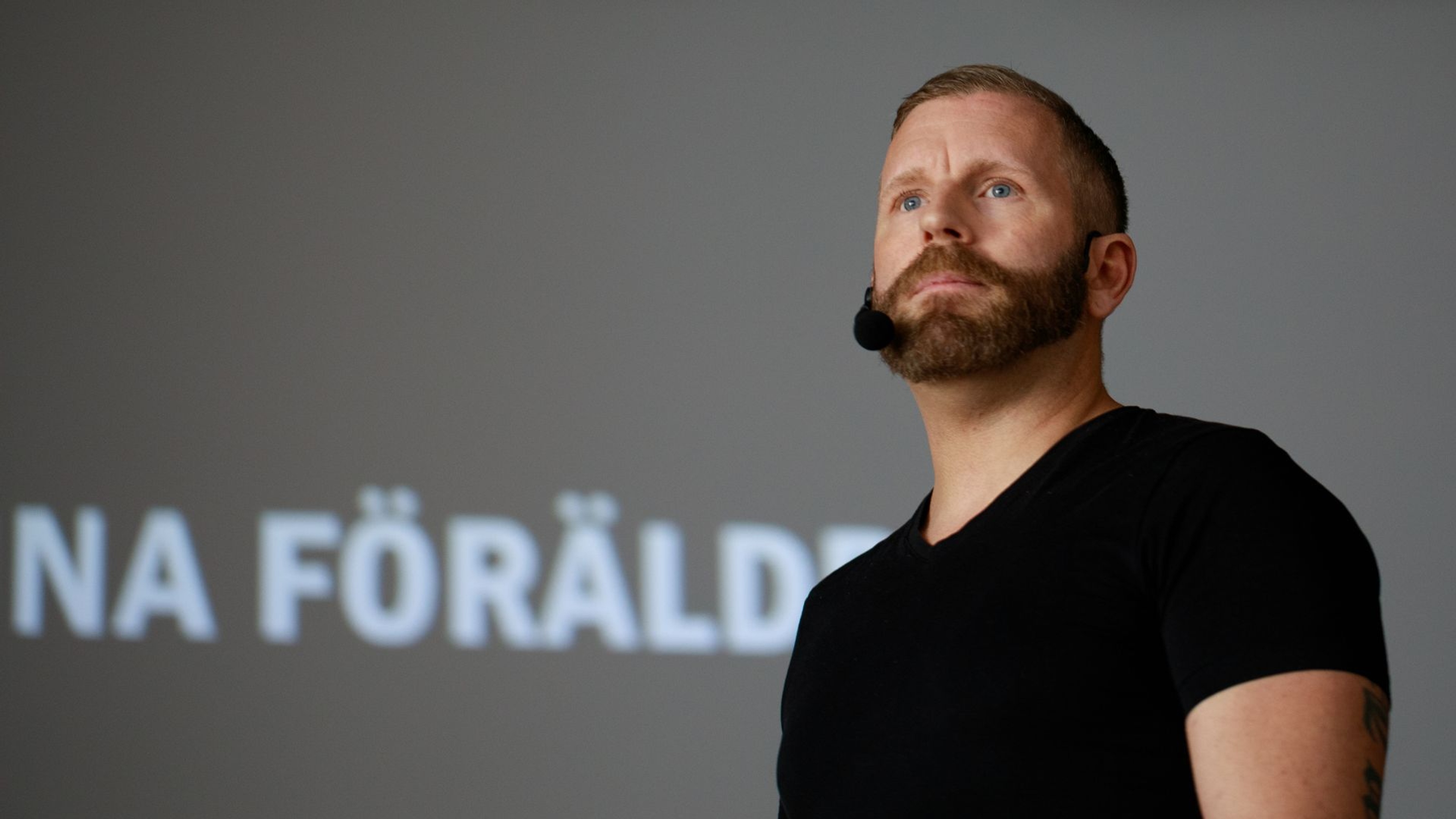 Stefan Carter, med svart t-shirt och headset framför en grå bakgrund med suddig text.