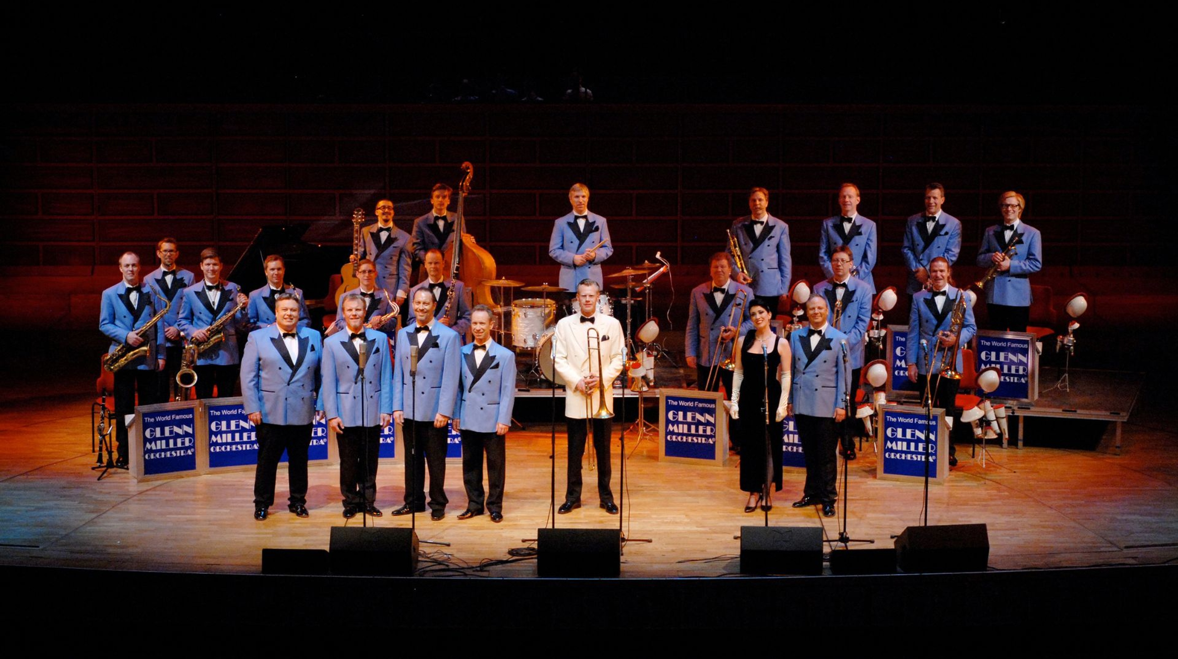 Orkestern står uppställda på scen iklädda blå kostymer. 