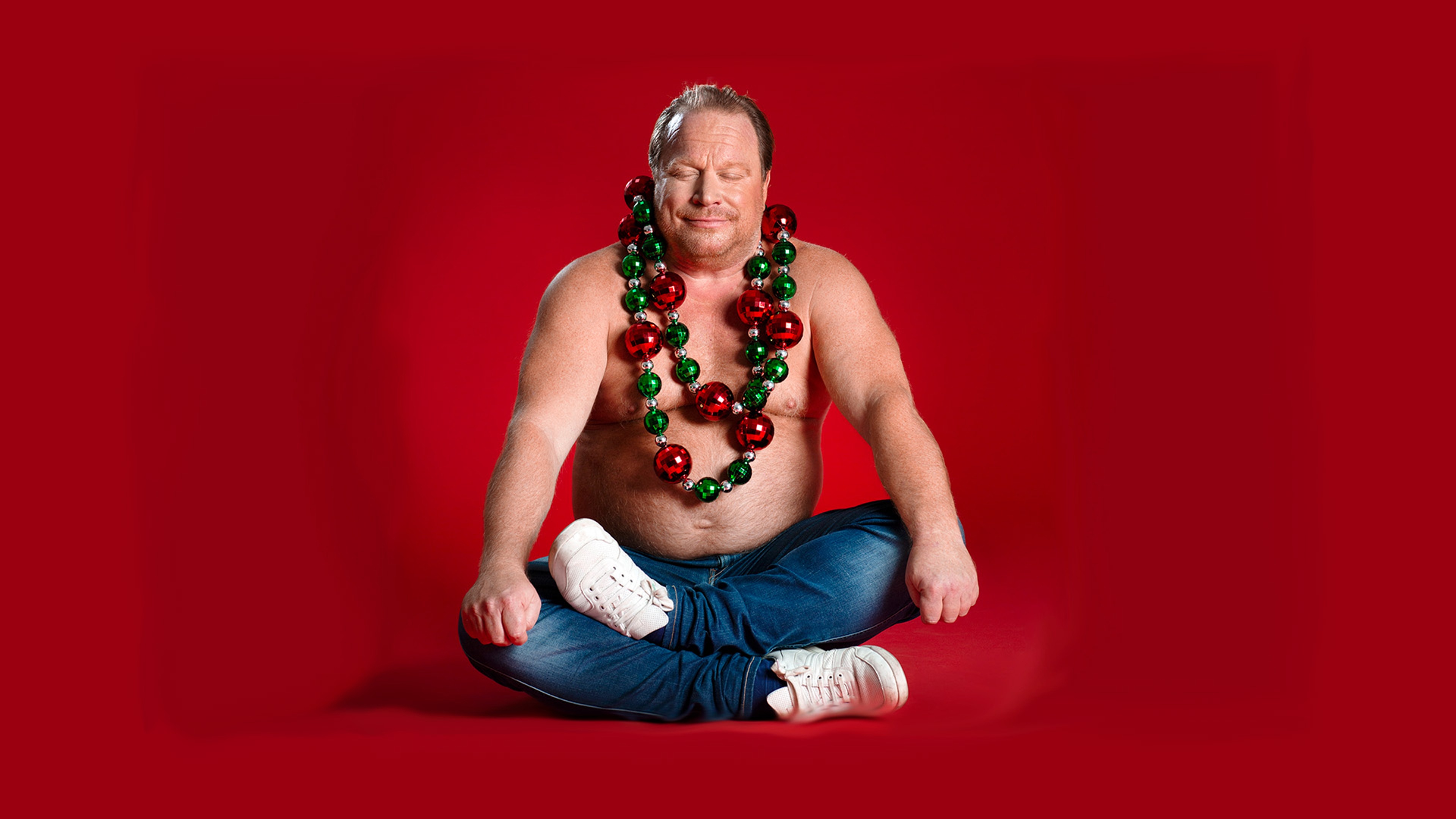 Claes Malmberg sitter skräddare med bar överkropp och röda och gröna julkulor runt halsen. 