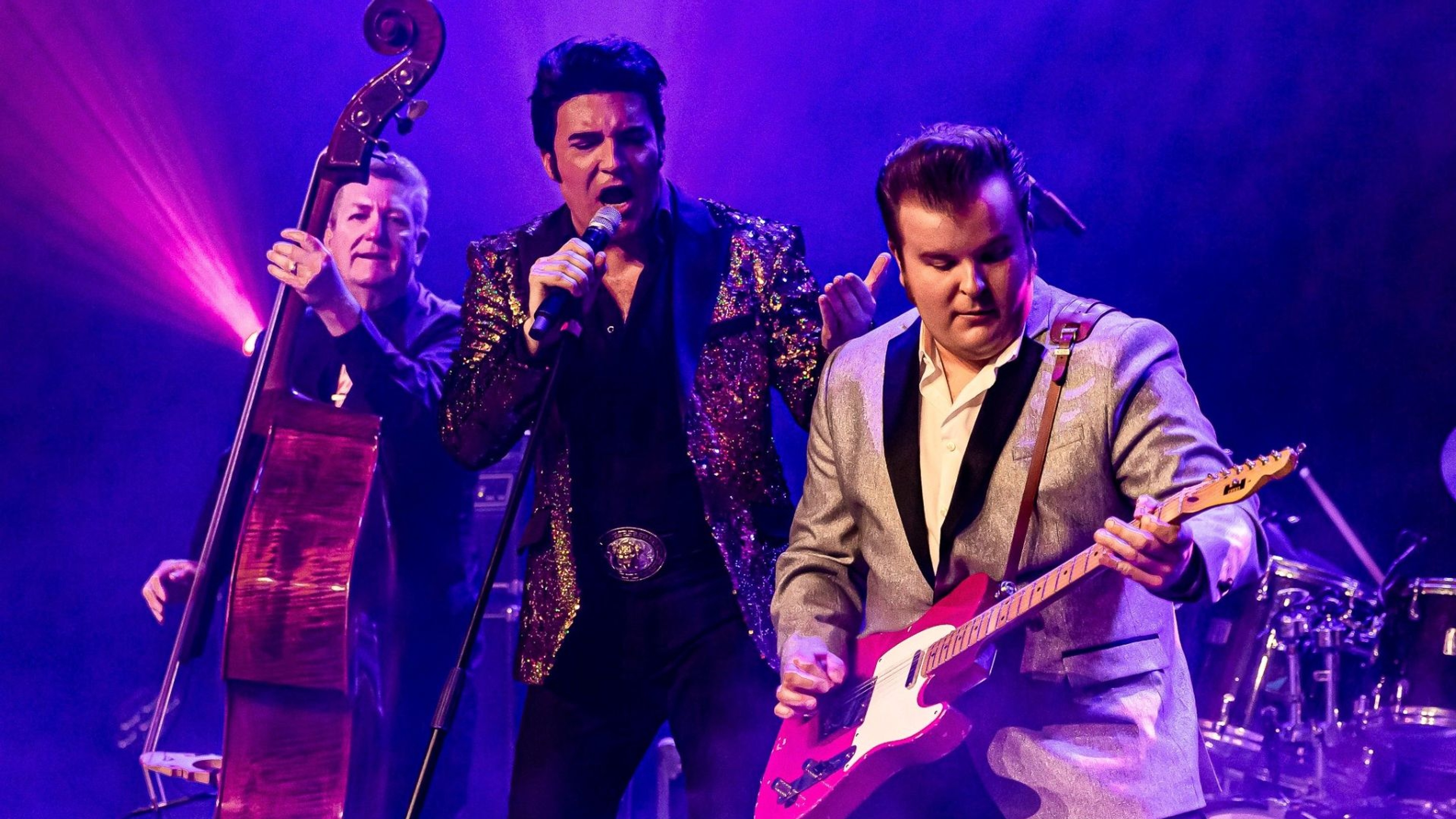 Dwight Icenhower som Elvis står i mitten och sjunger i mikrofon. Till vänster spelar en man kontrabas och till höger spelar en man elgitarr.