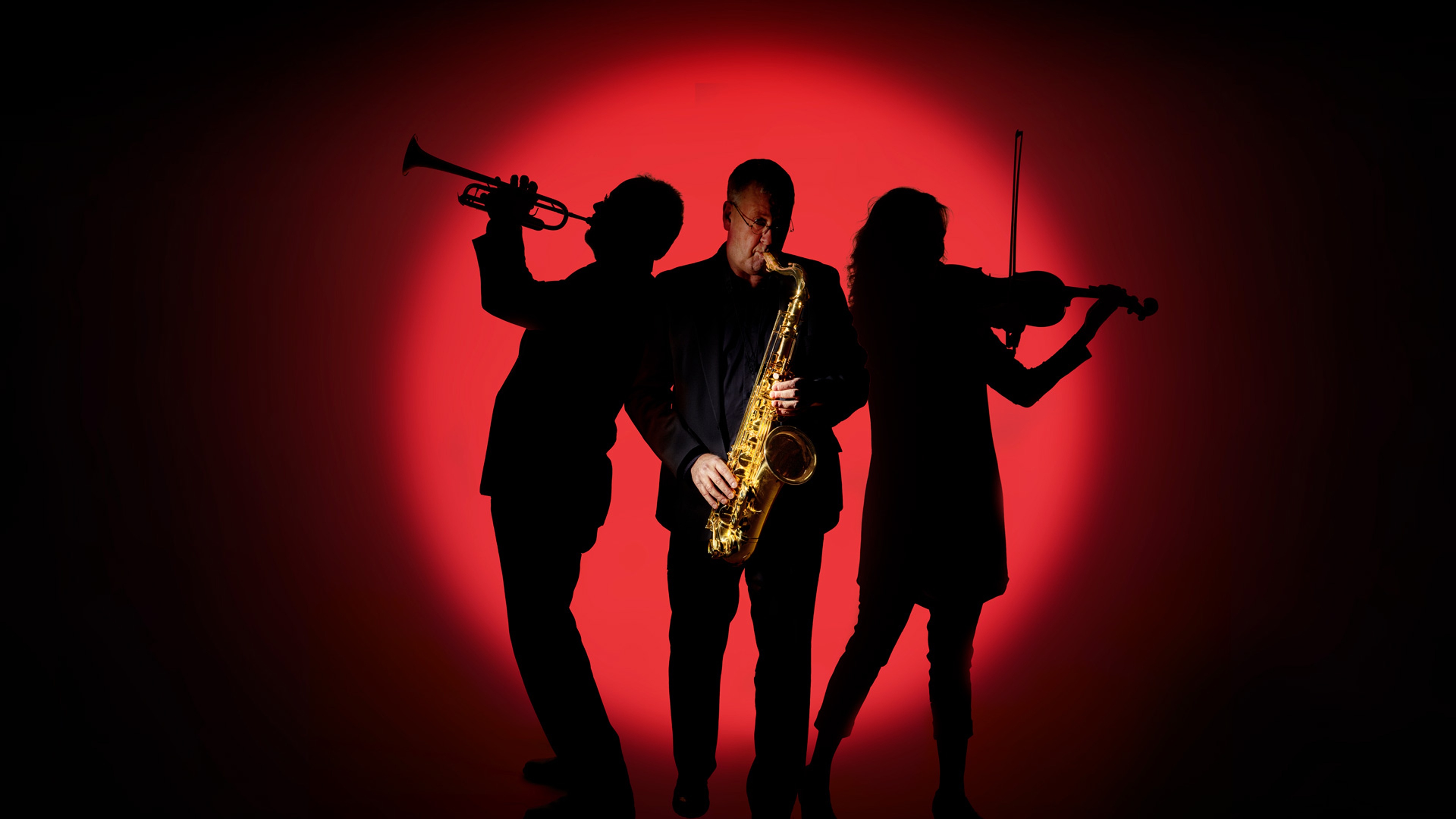 Tre musiker framför en röd bakgrund. En trumpetare till höger, en saxofonist i fokus i mitten och en violinist tillhöger.