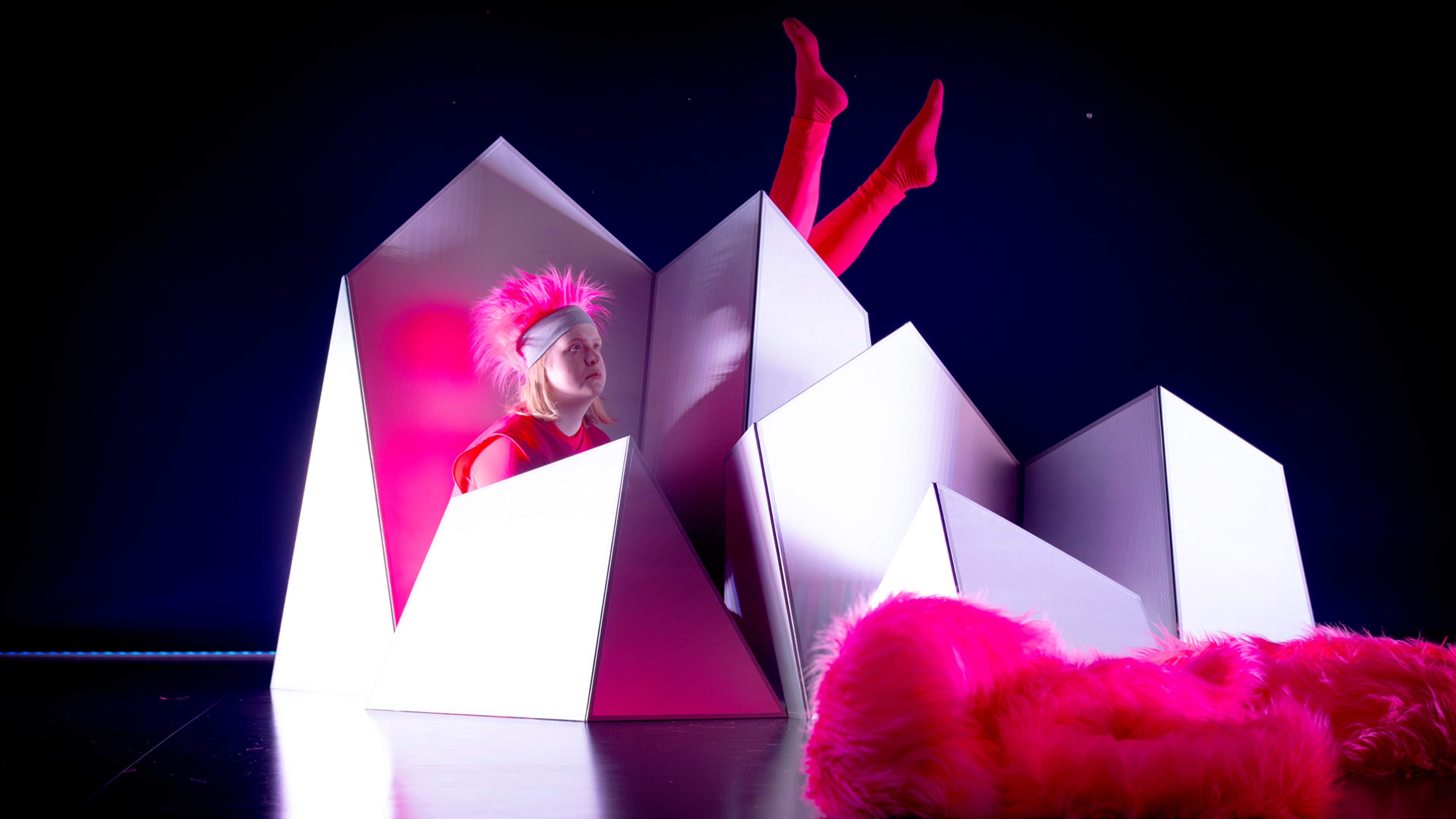 Vita boxar står på golvet, upp sticker ett par fötter iklätt rosa och en person med fluffig rosa huvudbonad.