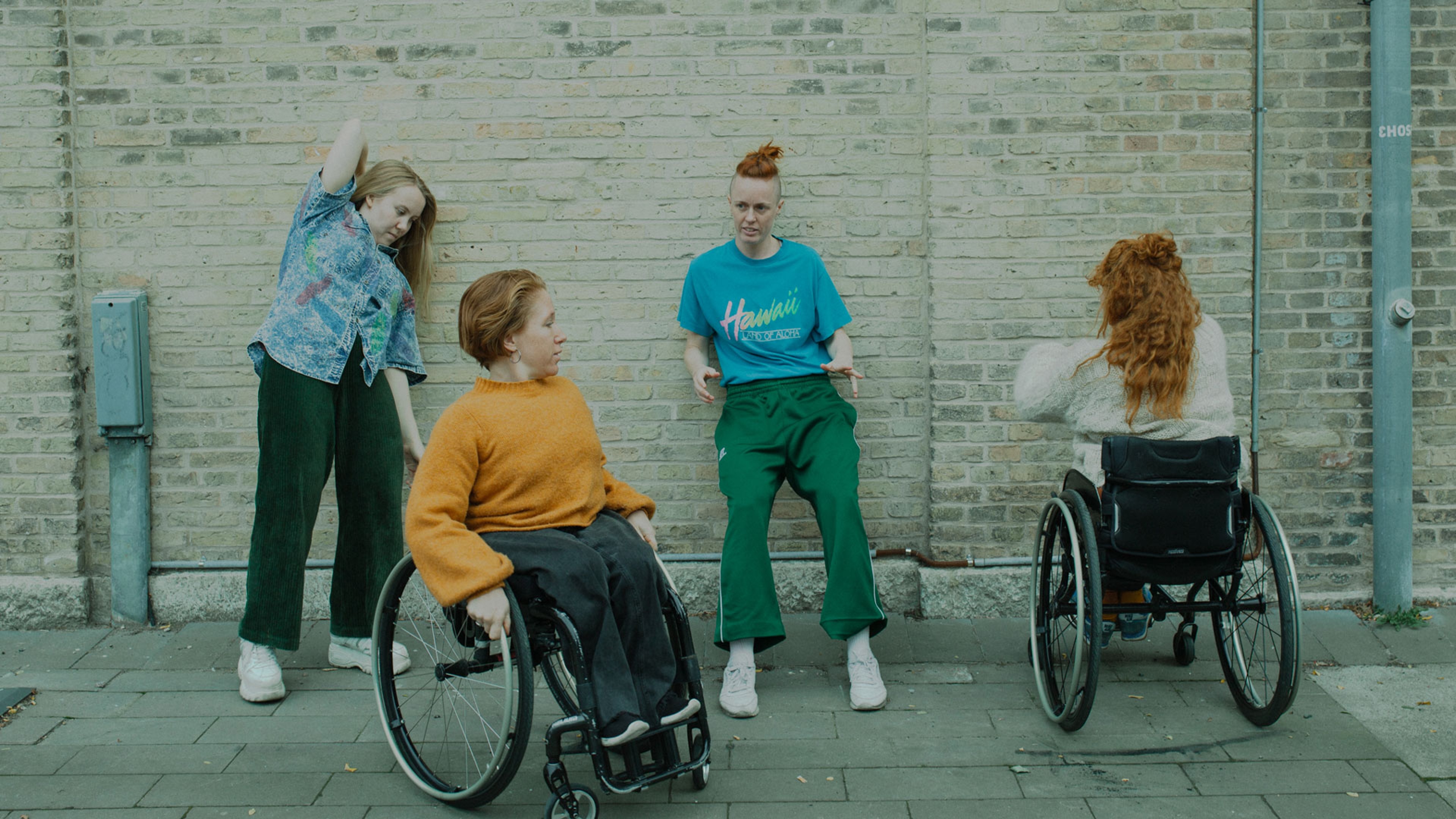 Fyra dansare mot en tegelvägg. Två står upp och två sitter i rullstol.