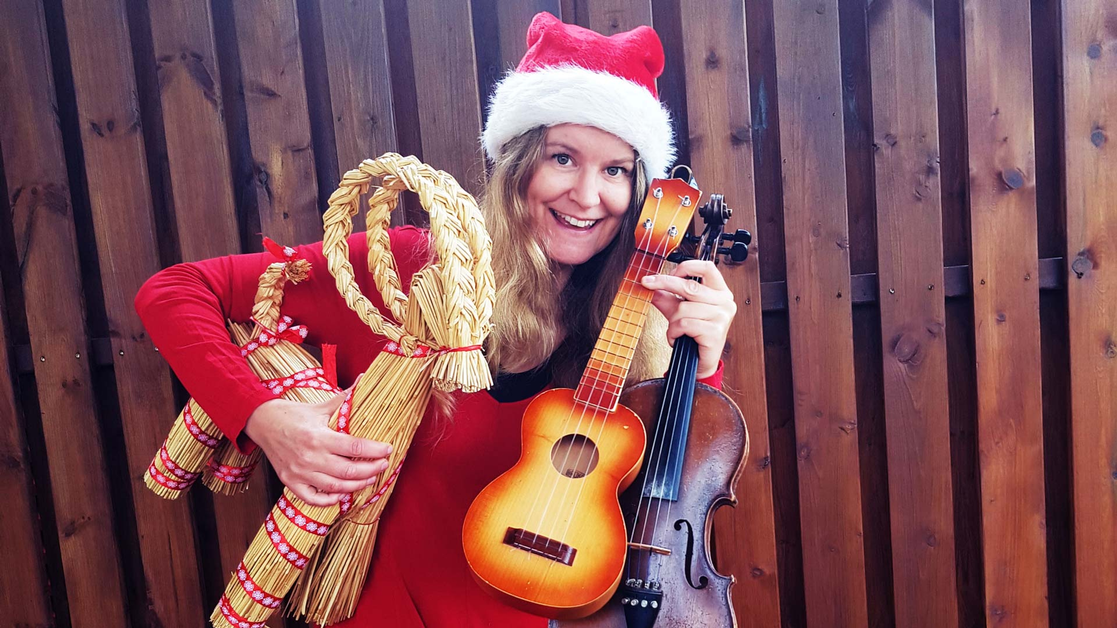 En leende Susanne Lind står mot ett träplank och håller i en julbock, ukulele och fiol. På huvudet har hon en tomteluva.