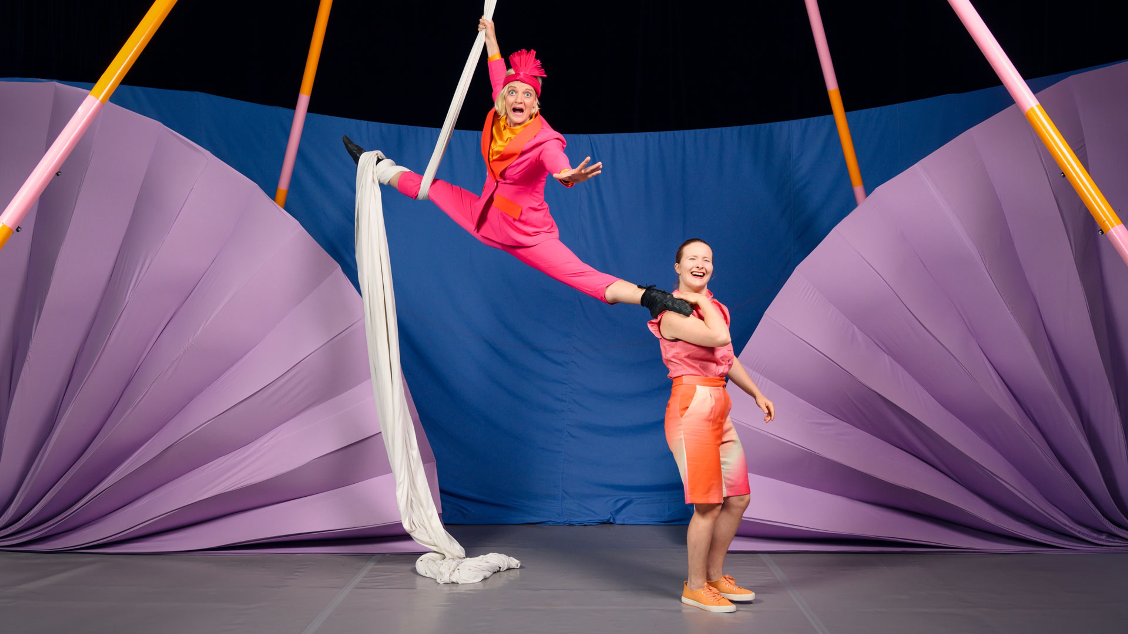Två cirkusartister varav den ena hänger ned från taket framför en vägg i blått och lila