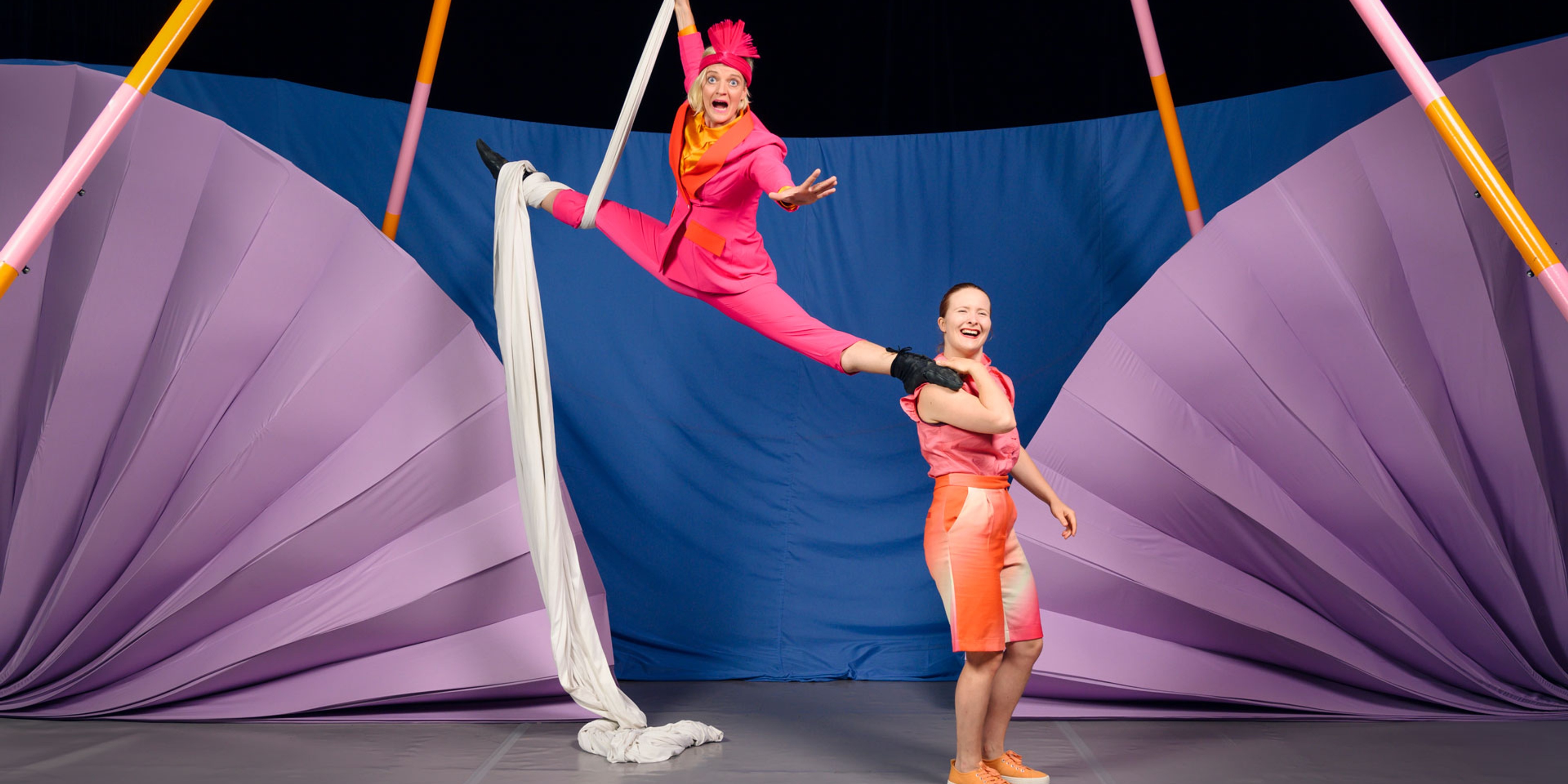 En cirkusartist hänger ned från taket och en står på marken med den hängande personens fot på sin axel. I bakgrund lila och blå bakgrund.