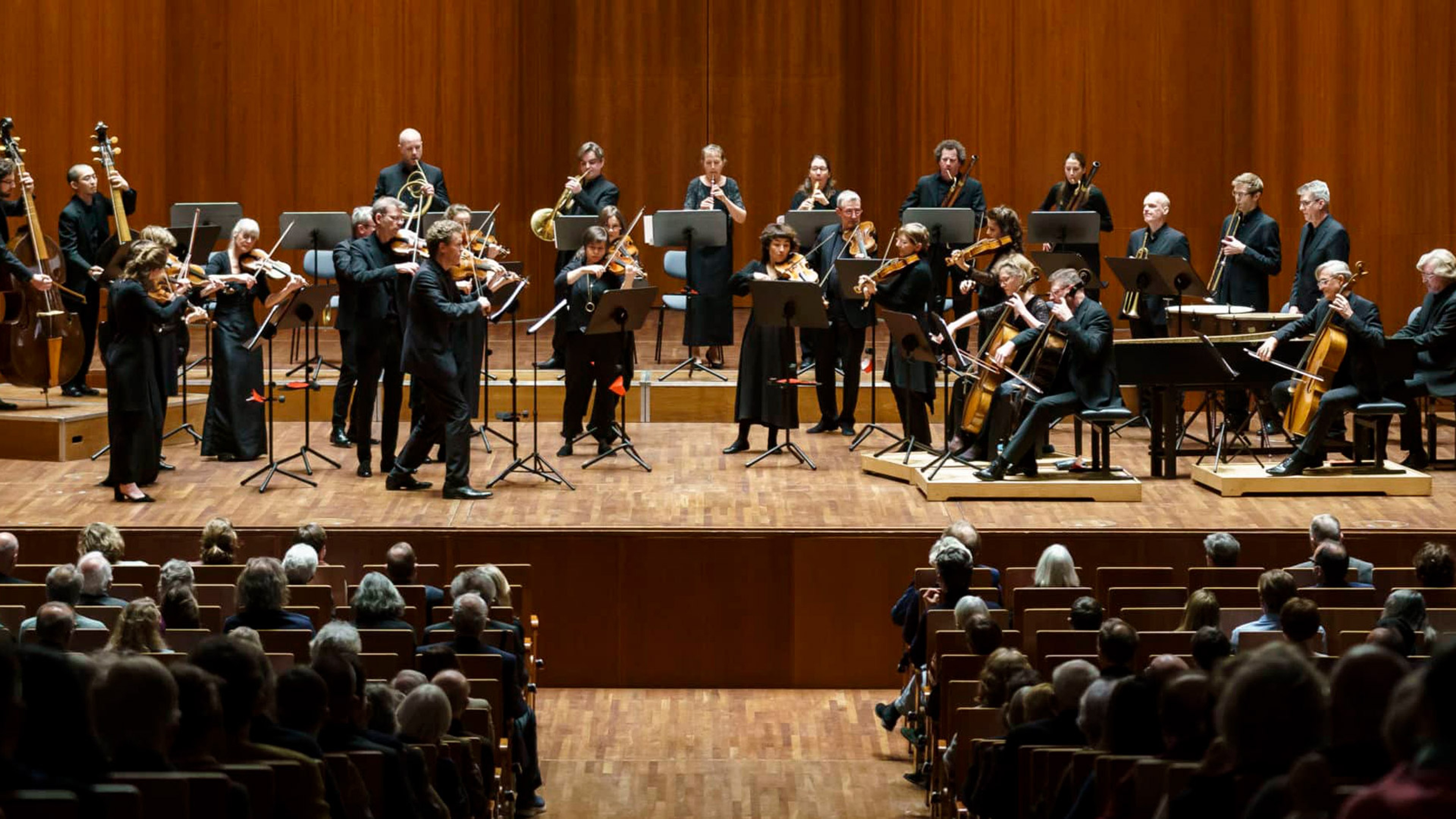 Orkester står på scenen med en orange bakgrund och framför en konsert med publik förgrunden