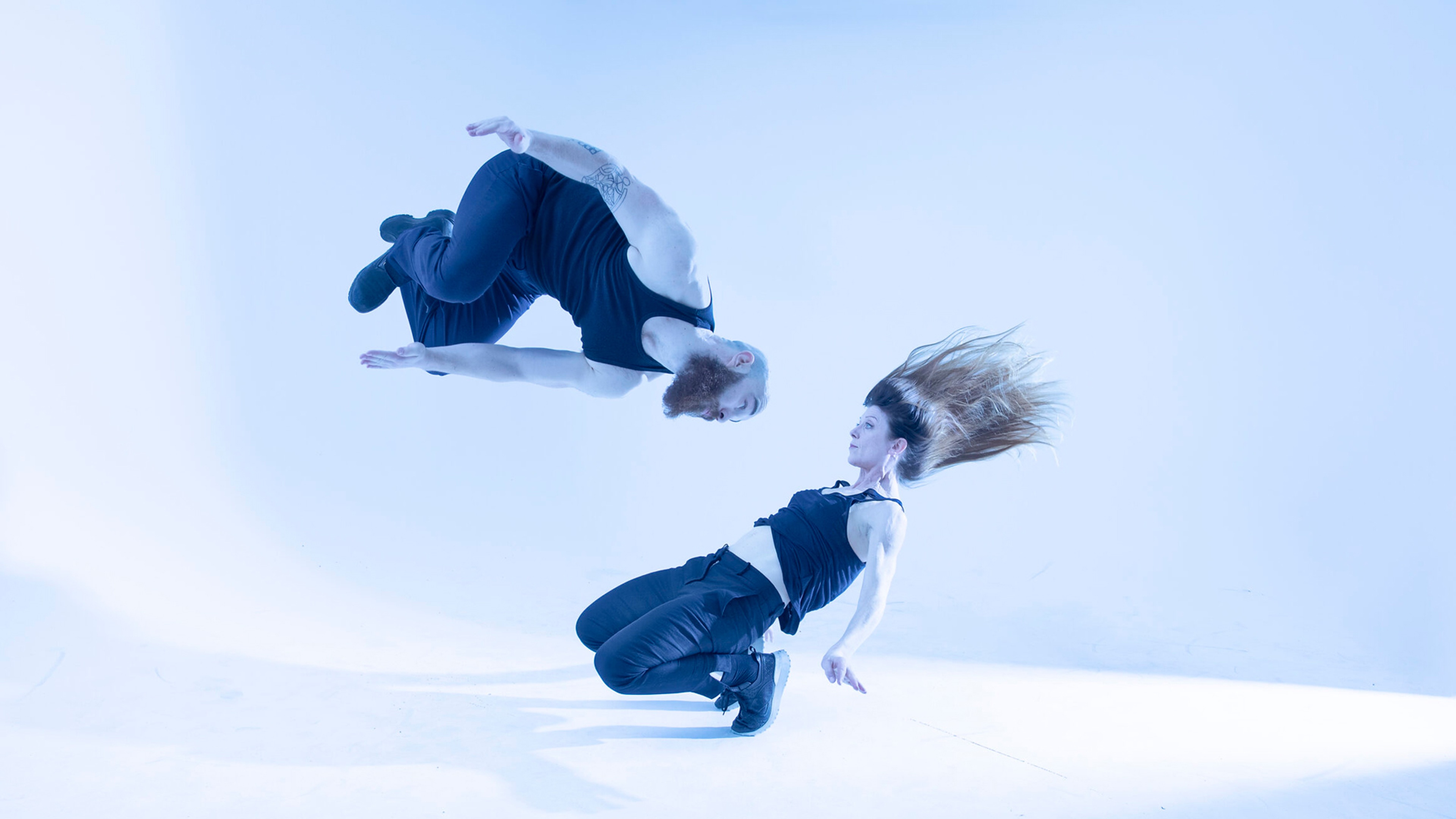Två dansare i rörelse, en ner mot golvet och en i luften i en volt.