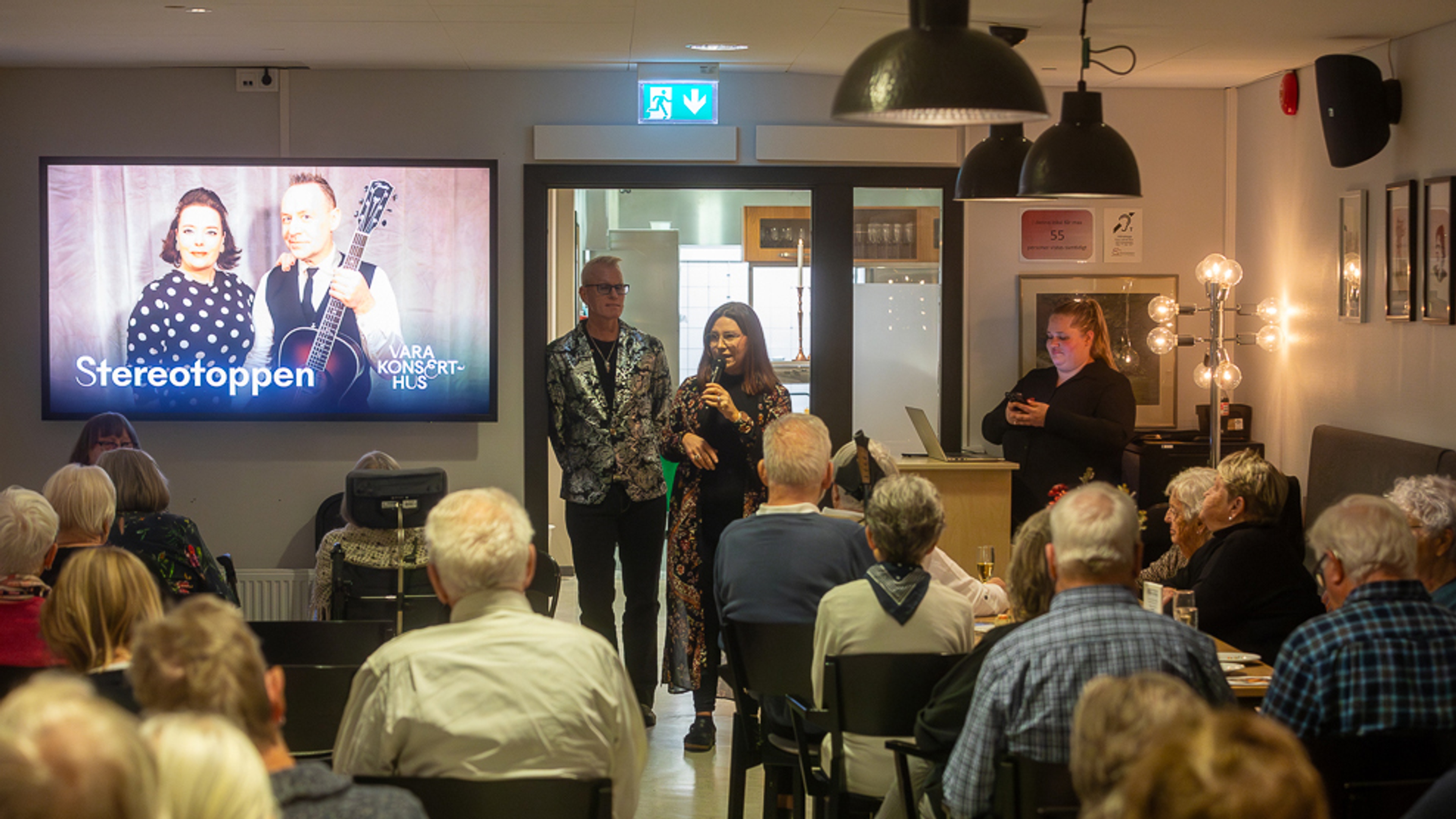 Publik och en skärm med scenkonst. En person håller en introduktion.